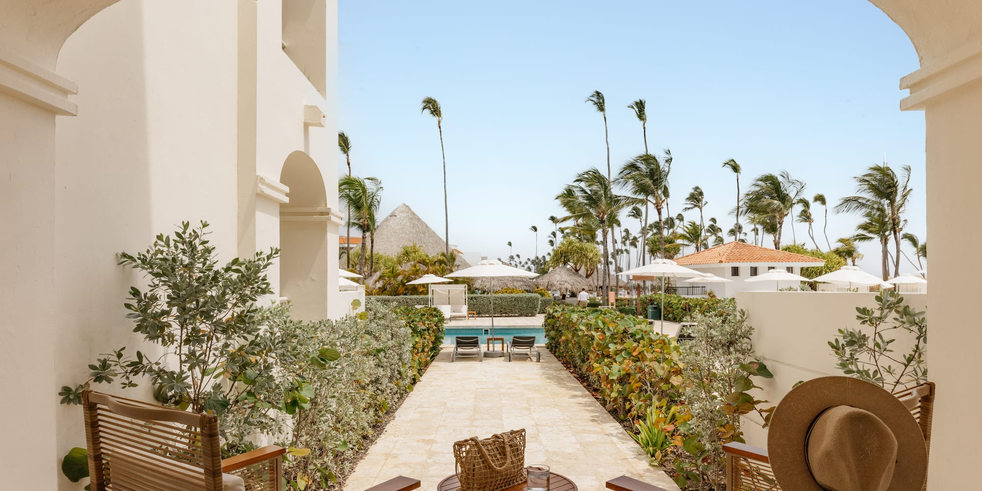 Arched patio overlooking tropical resort pool, swaying palm trees & cabanas.