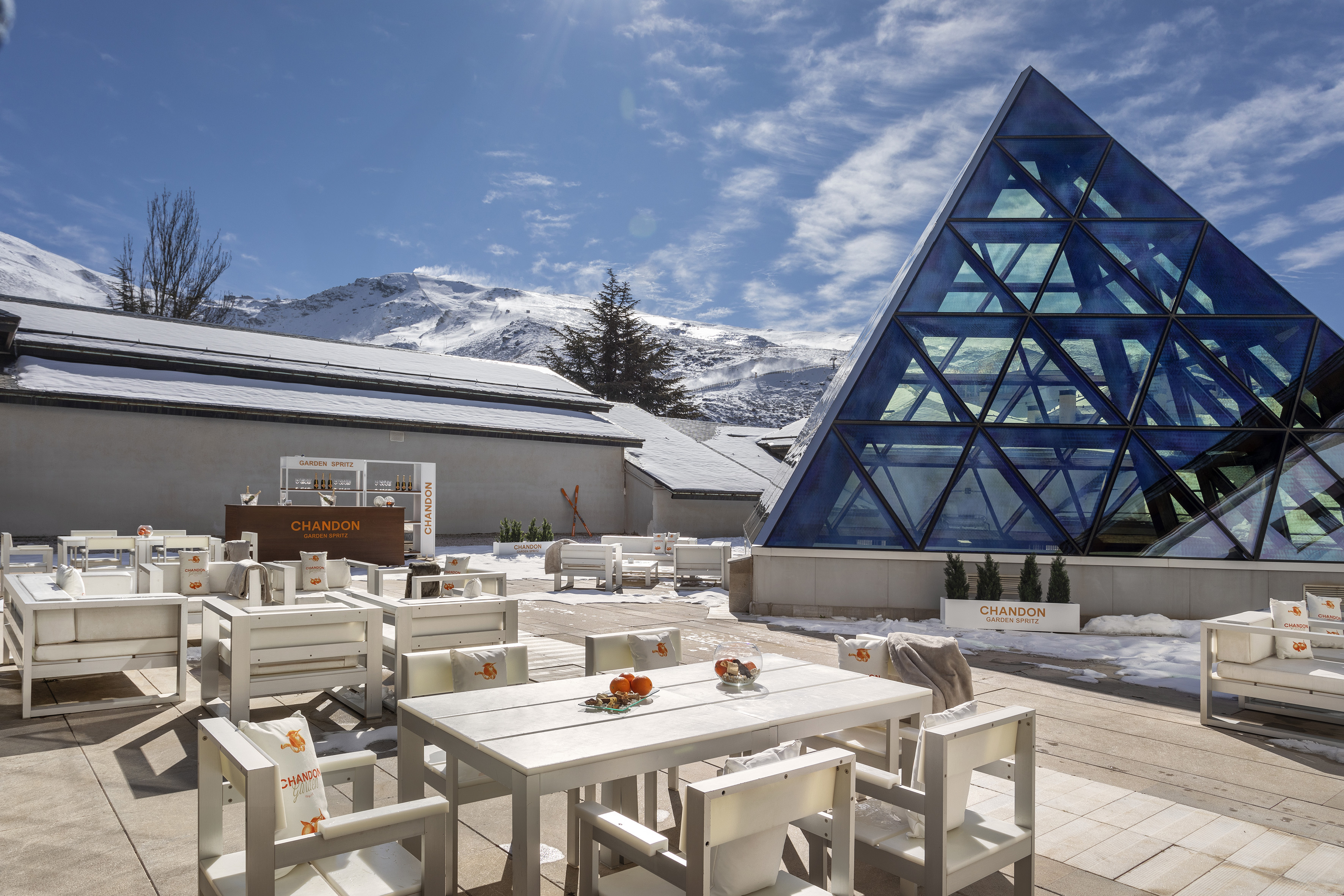 Luxury mountain resort patio with white furniture, modern glass pyramid, and snowy peaks.