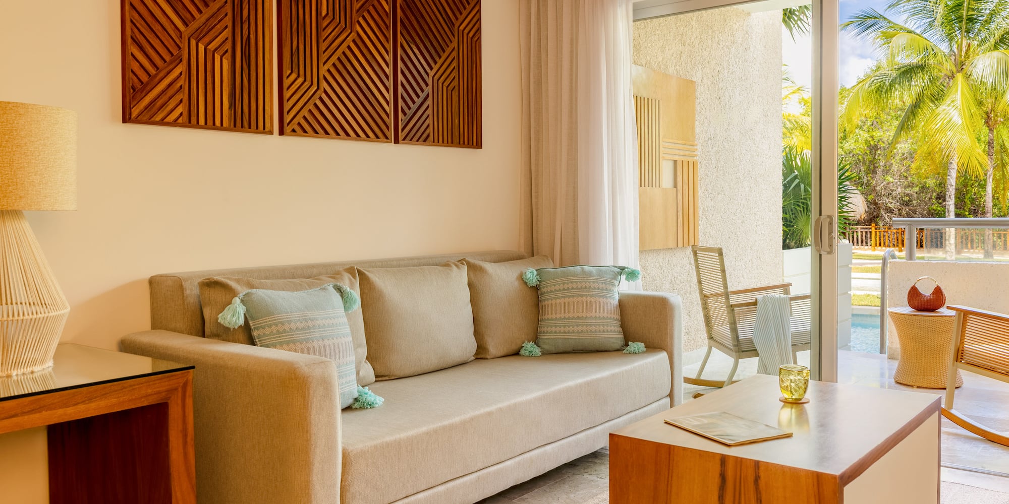 Tropical resort living room with beige sofa, wooden art, and patio pool view.