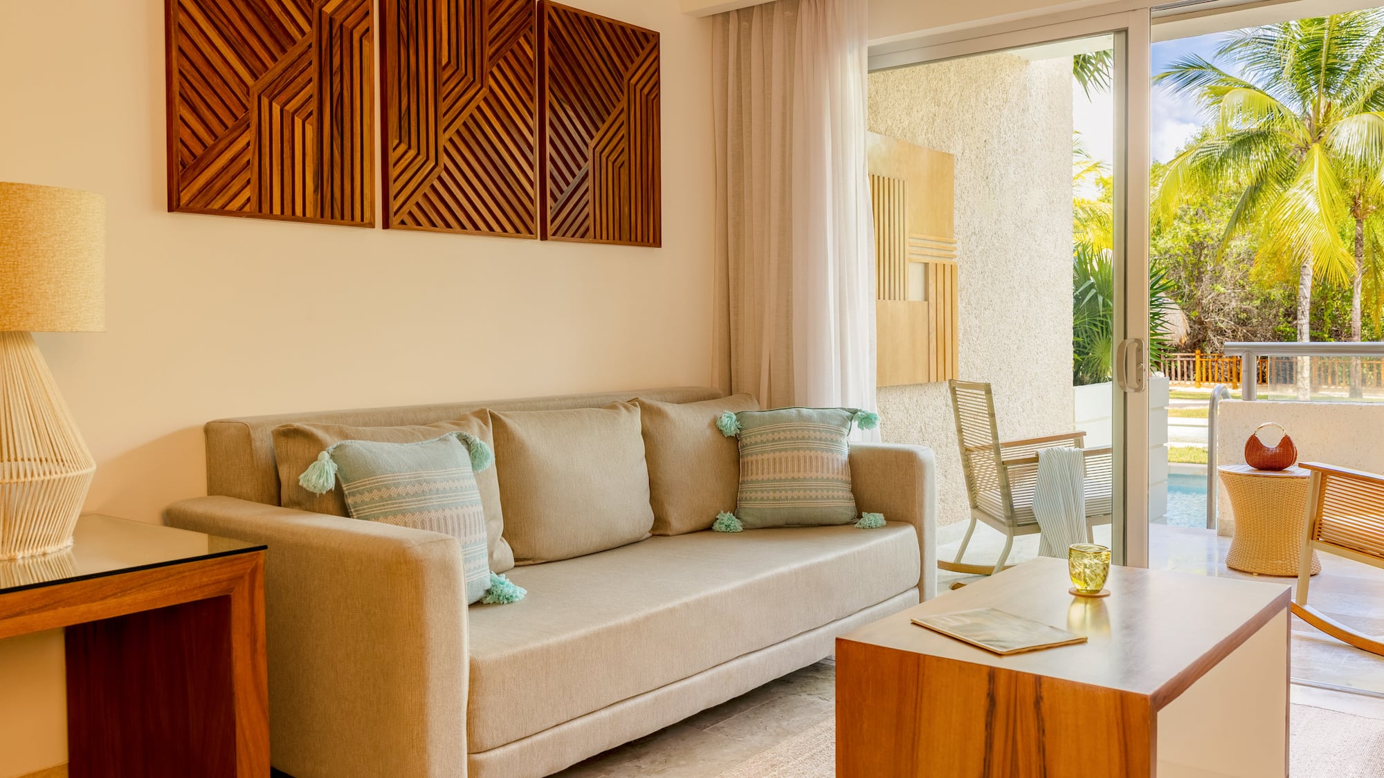Tropical resort living room with beige sofa, wooden art, and patio pool view.