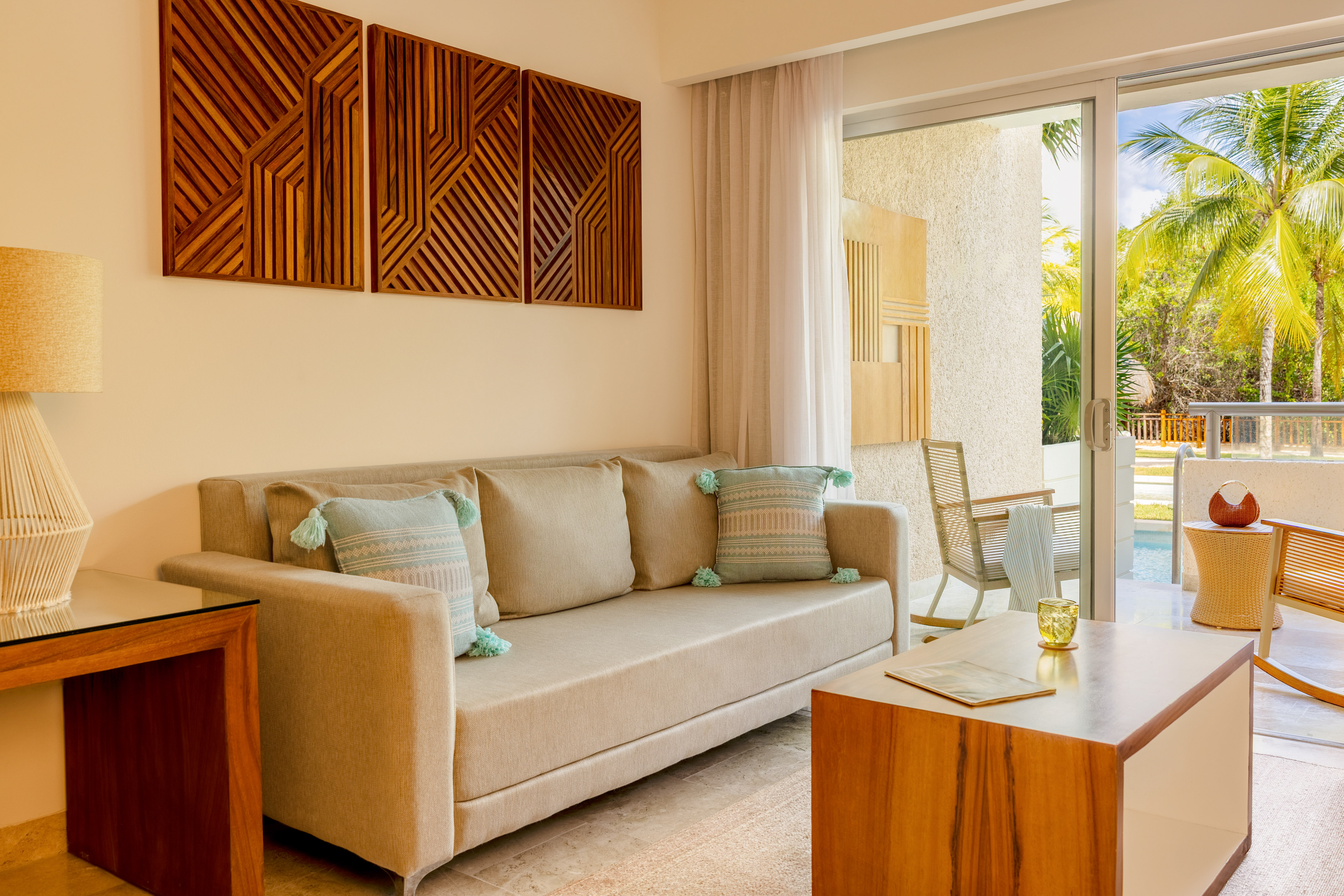 Tropical resort living room with beige sofa, wooden art, and patio pool view.