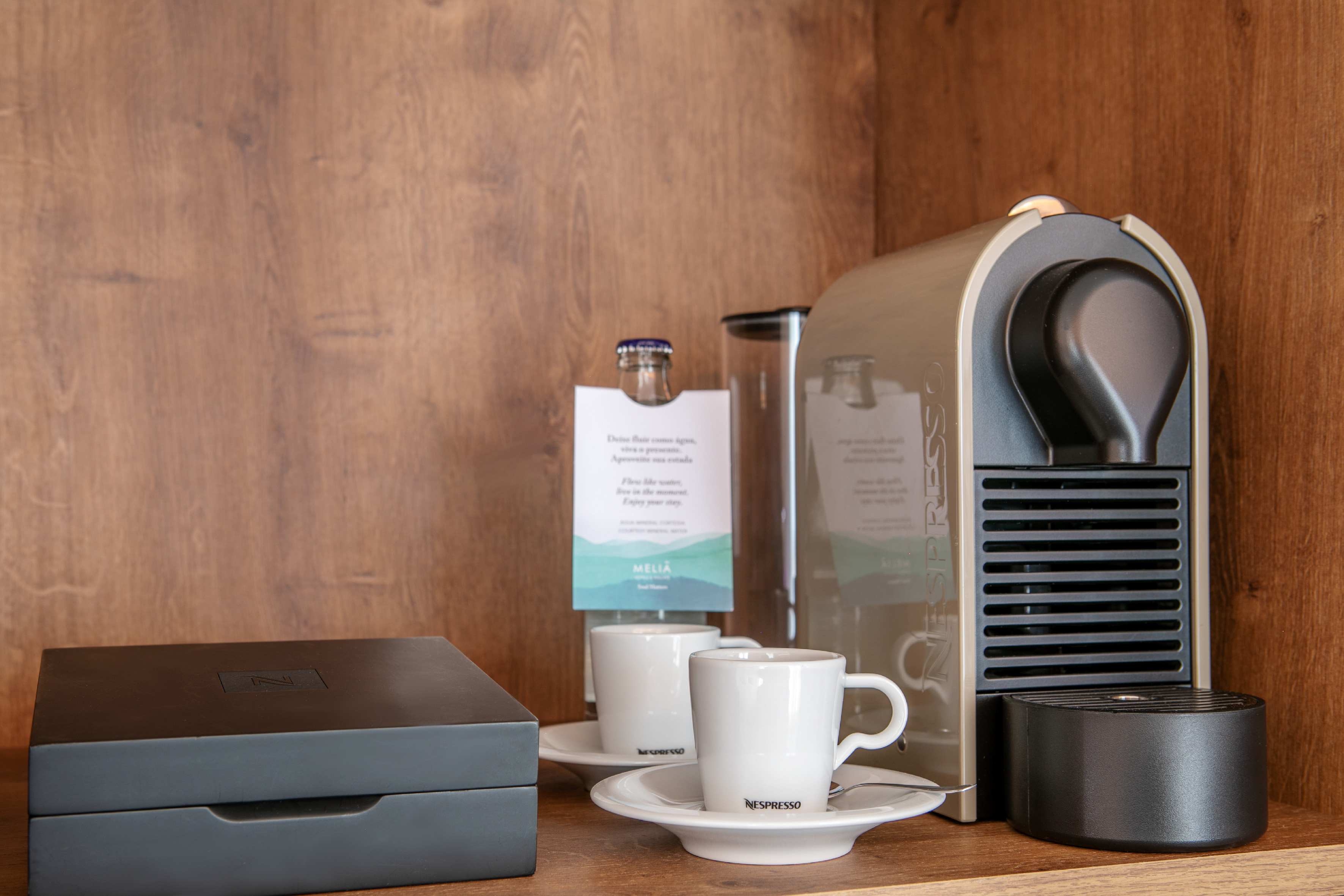 Hotel Nespresso coffee machine with cups, pod box, and water bottle on a shelf.
