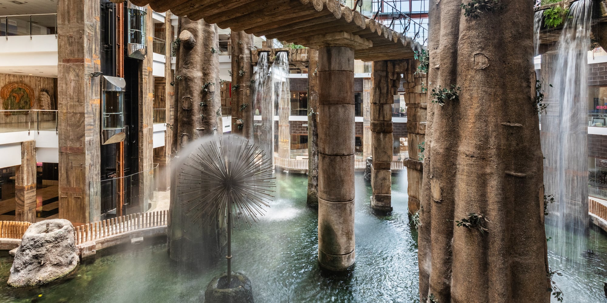 Indoor waterfall atrium with tree columns, log bridge, and starburst fountain.