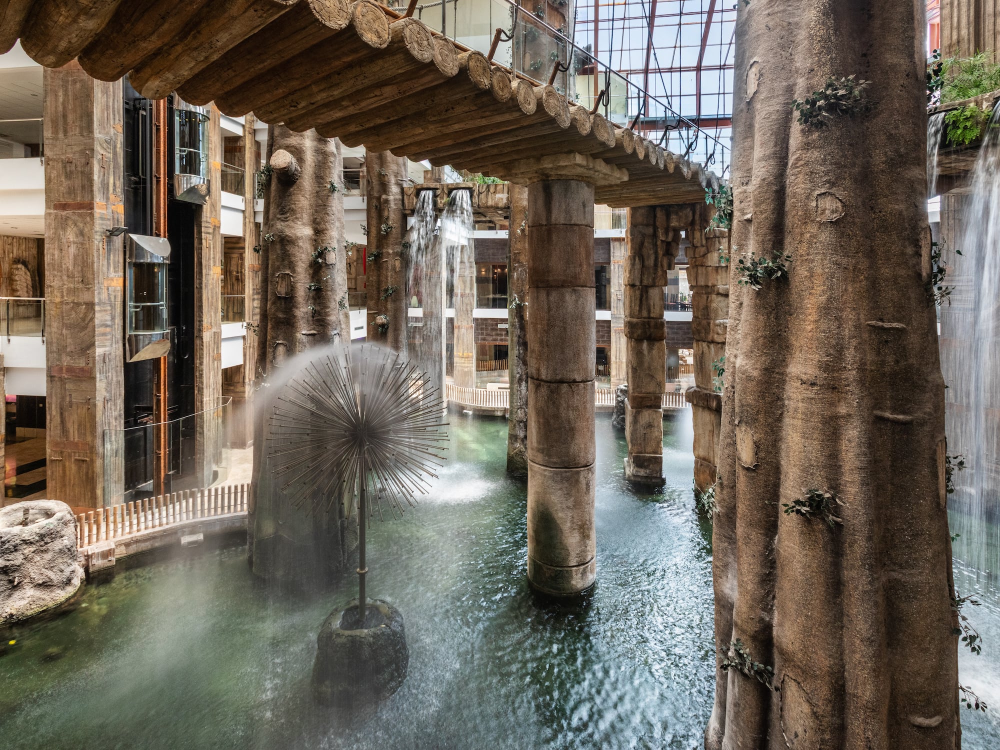 Indoor mall with waterfalls, artificial trees, fountain, and overhead bridge.