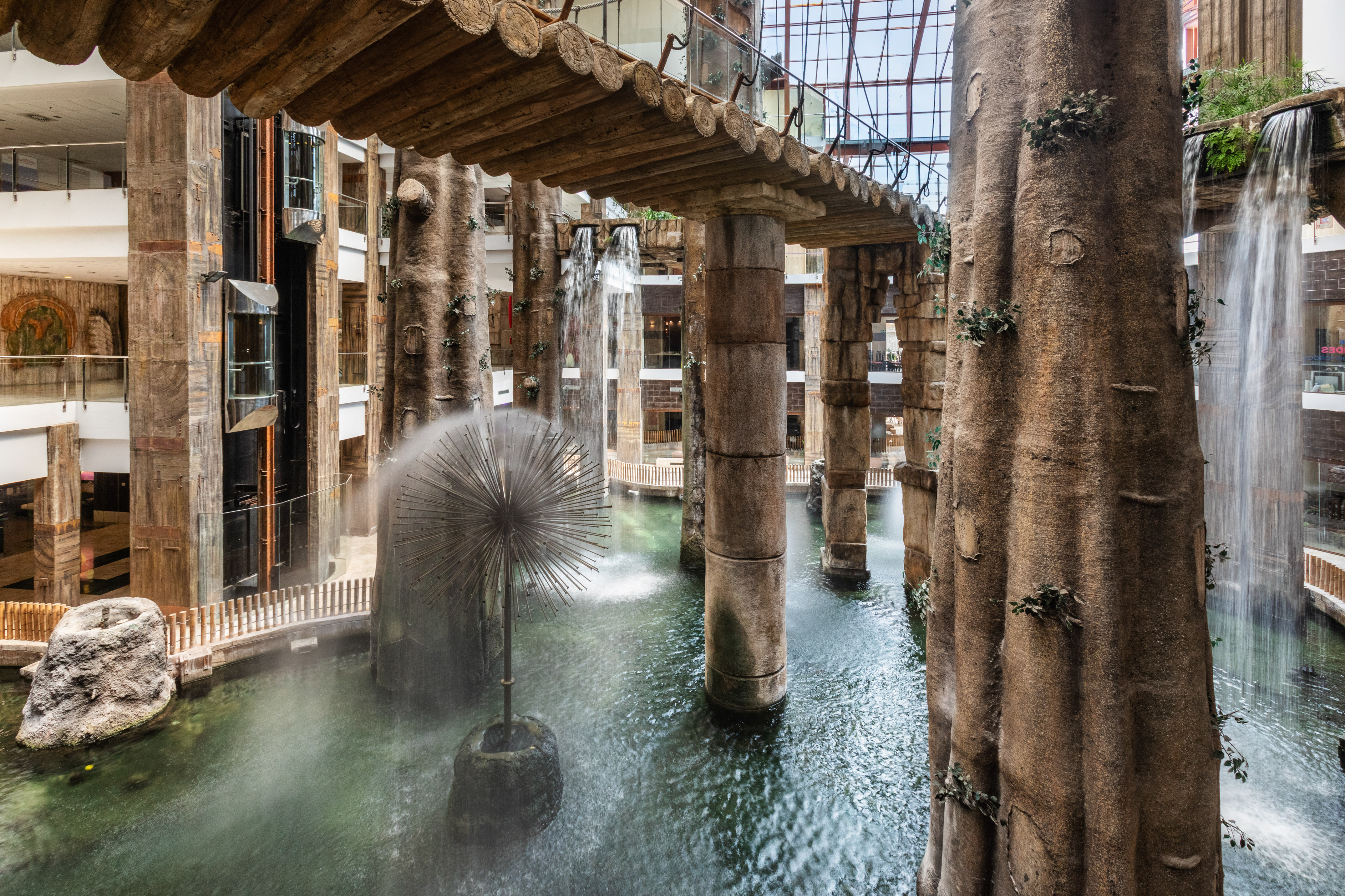 Indoor waterfall atrium with tree columns, log bridge, and starburst fountain.