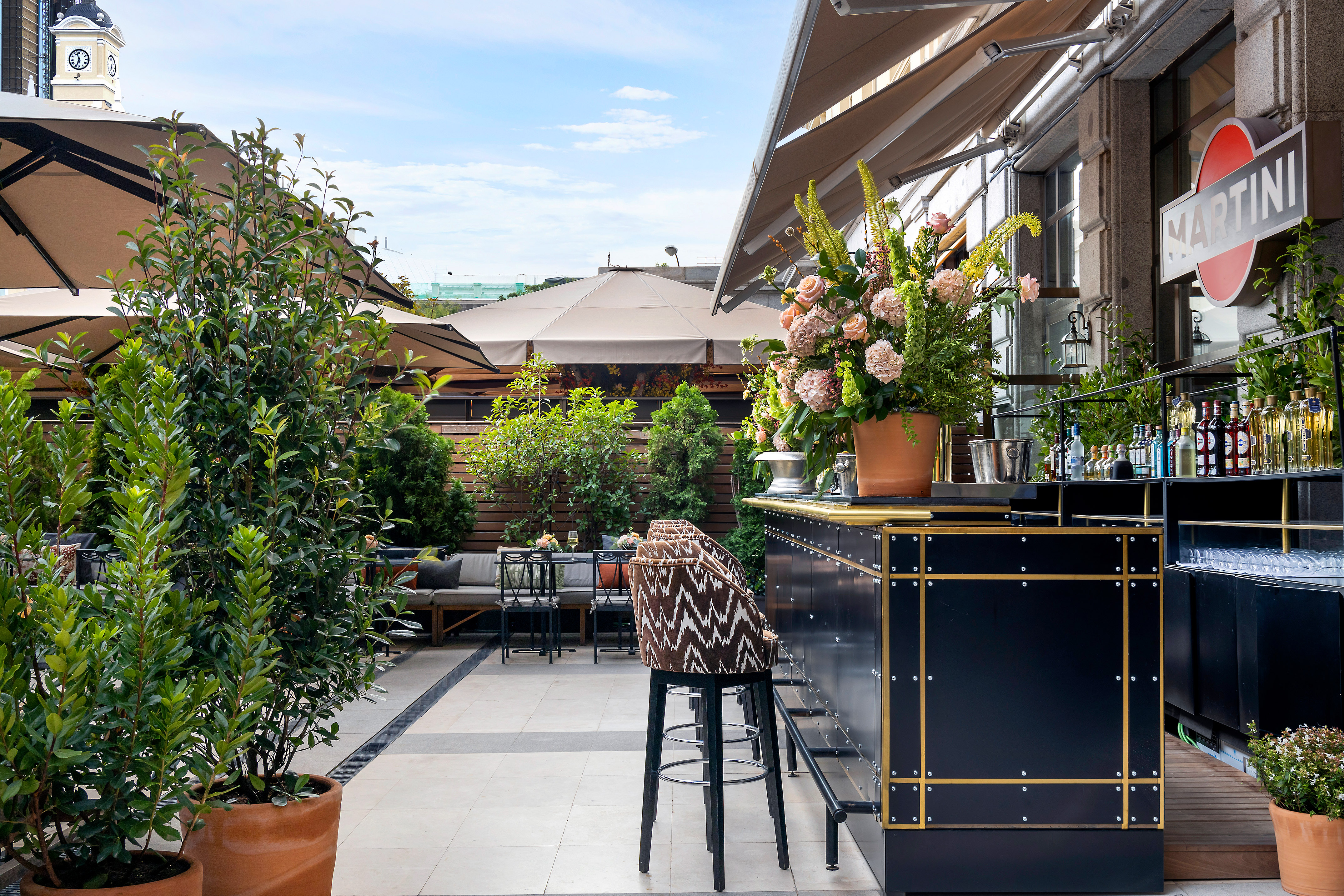 Rooftop bar with Martini sign, lush potted plants, and stylish outdoor seating area.