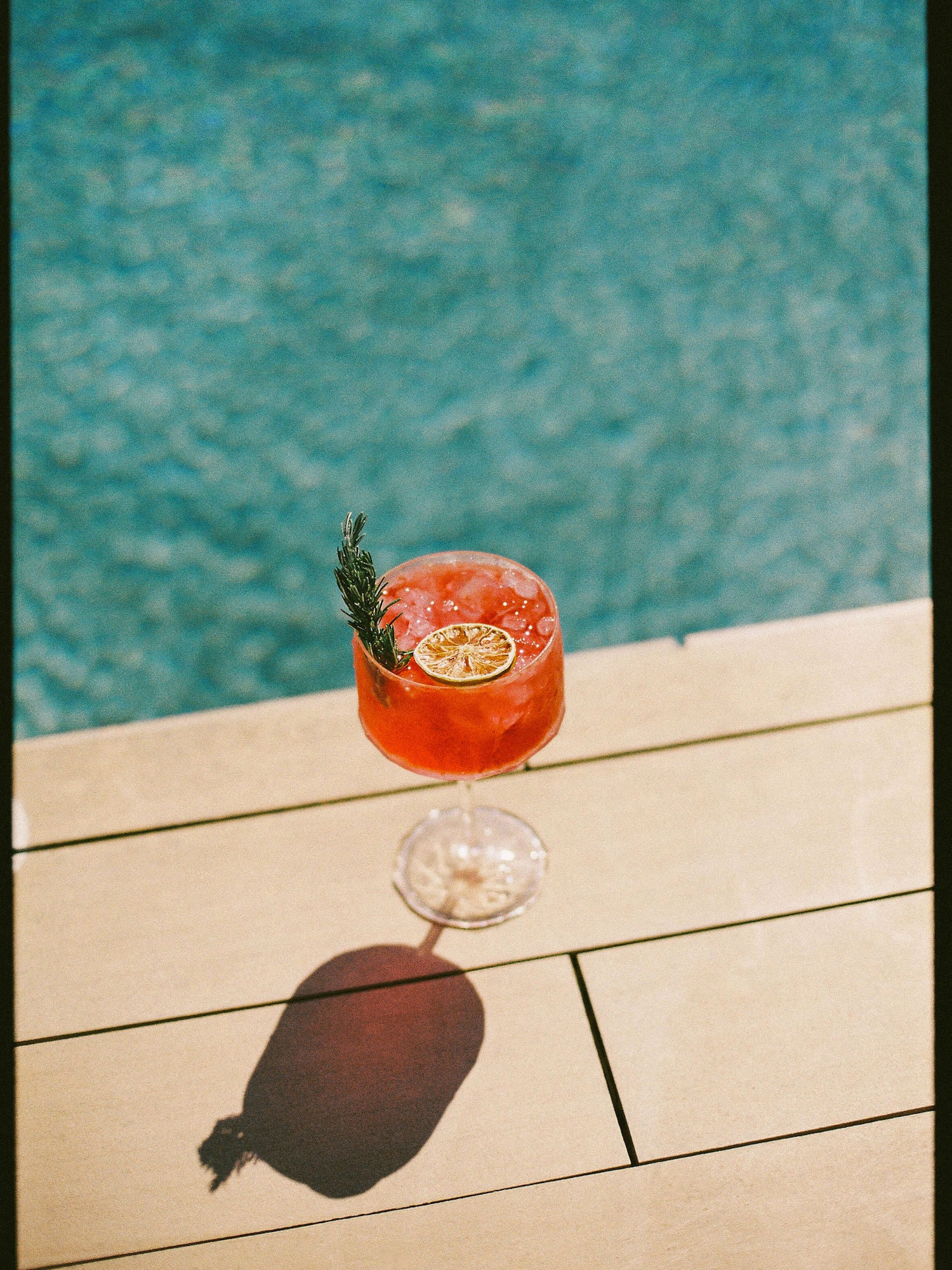 Red cocktail with rosemary & dried lime by a sunny pool.