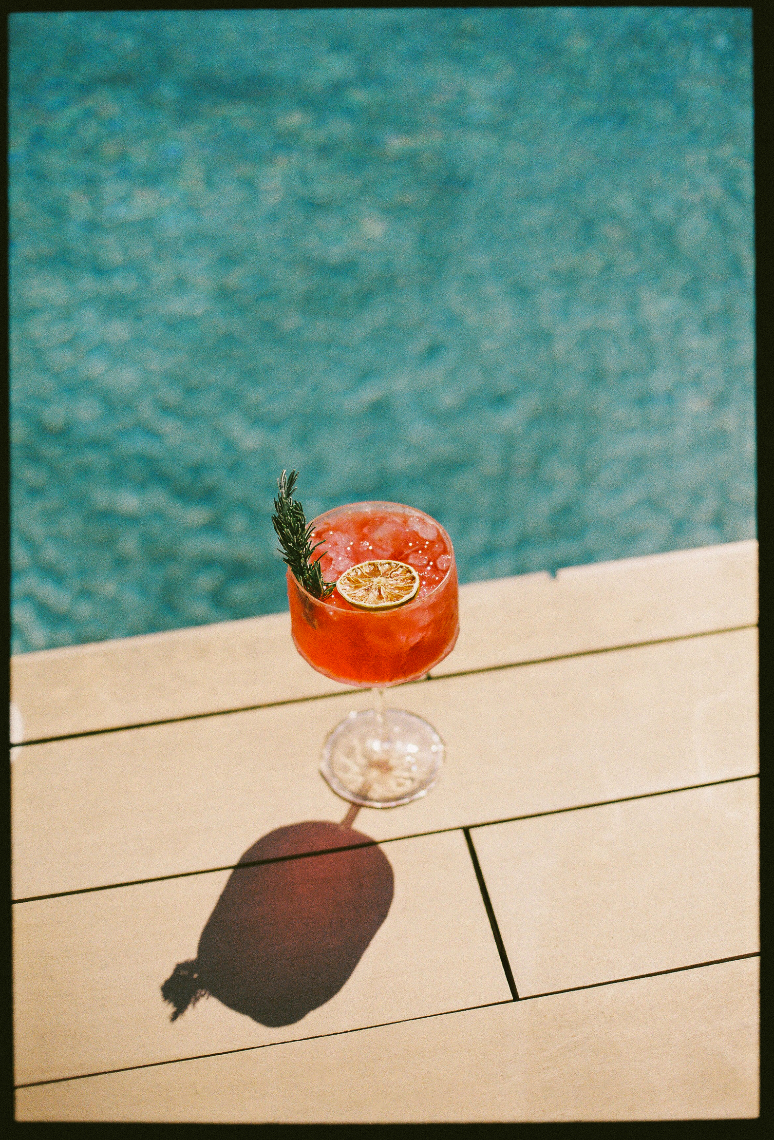 Red cocktail with rosemary & dried lime by a sunny pool.