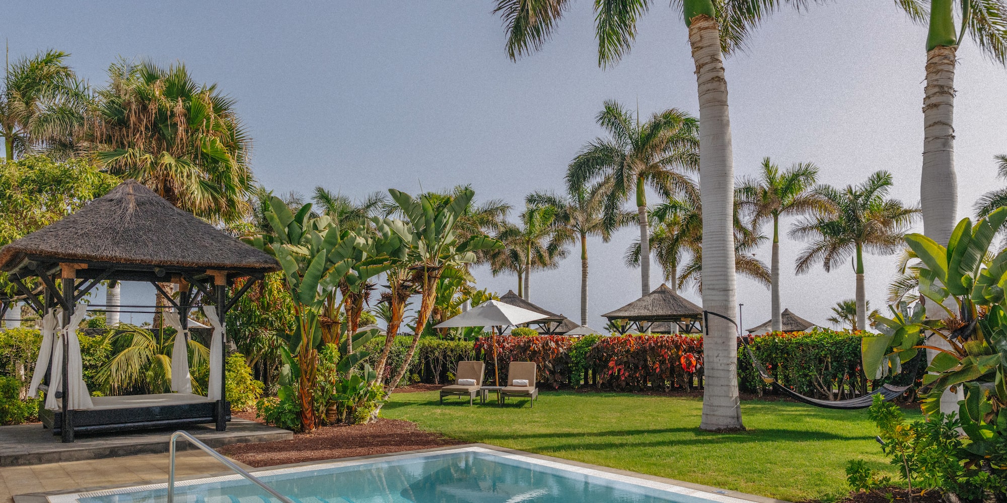 Lush tropical resort pool, thatched cabana, and tall palm trees.