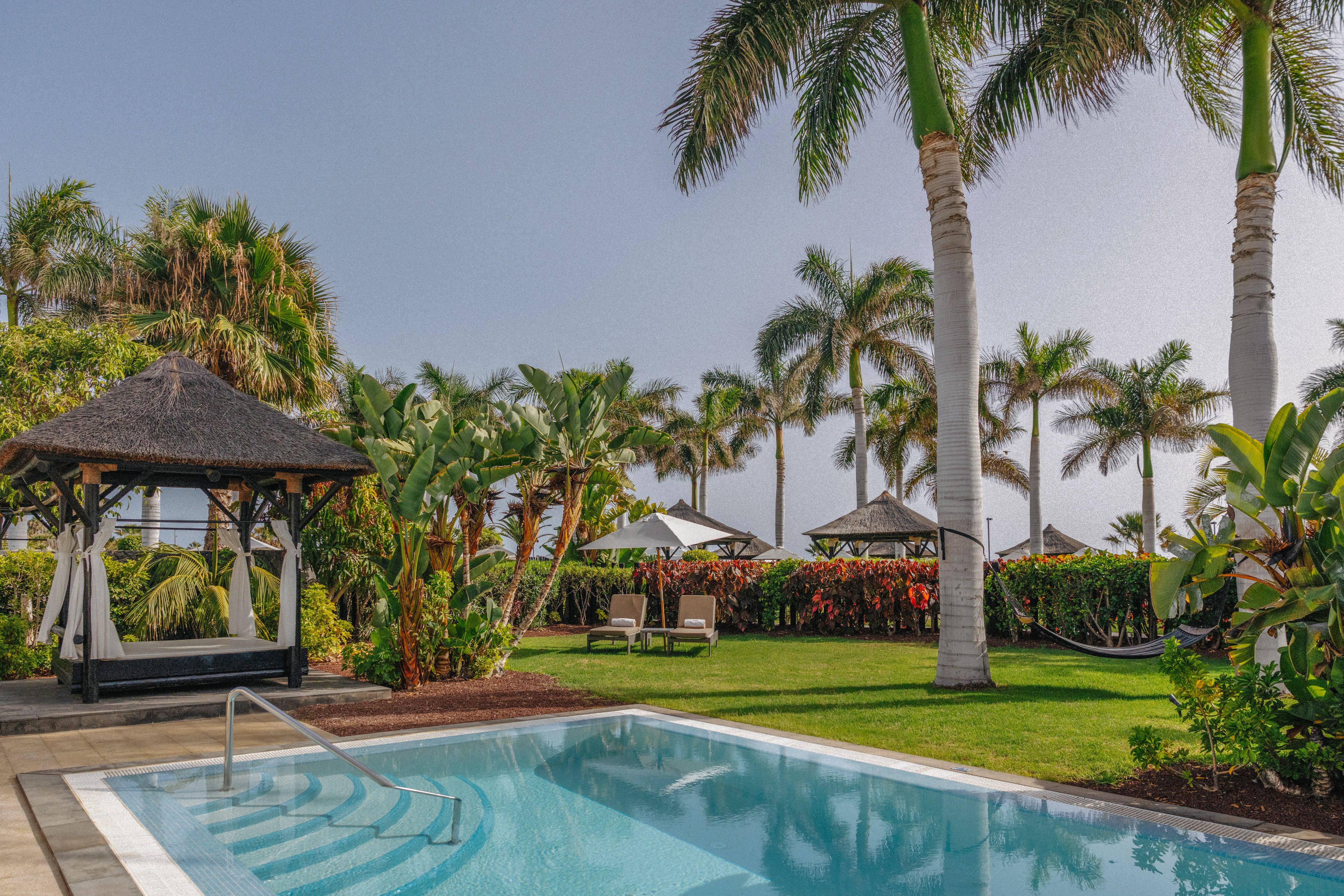 Luxury hotel private pool with tropical garden, palm trees, and thatched gazebo daybed.