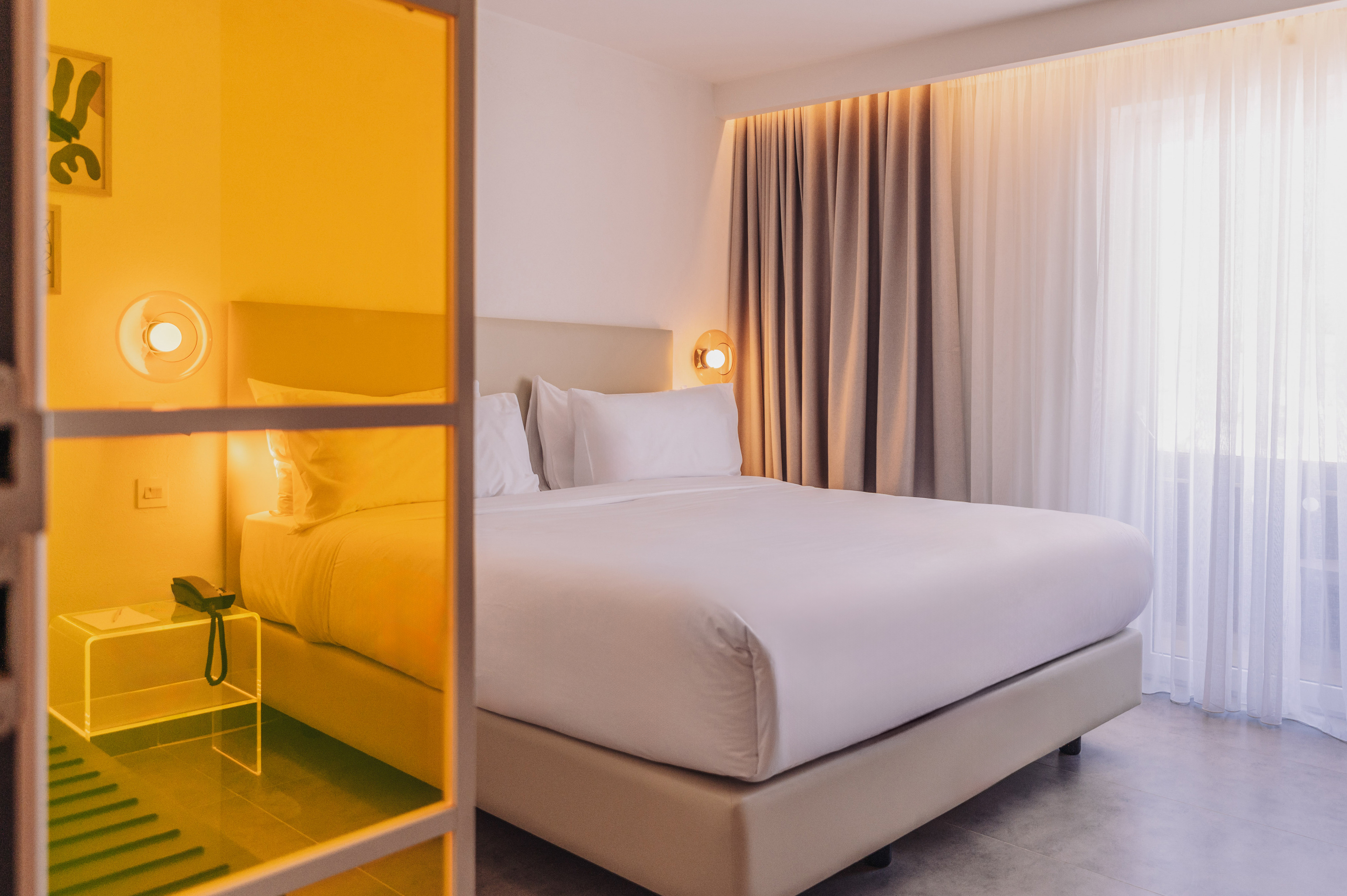 Modern hotel bedroom with king bed, neutral curtains, and yellow glass partition.