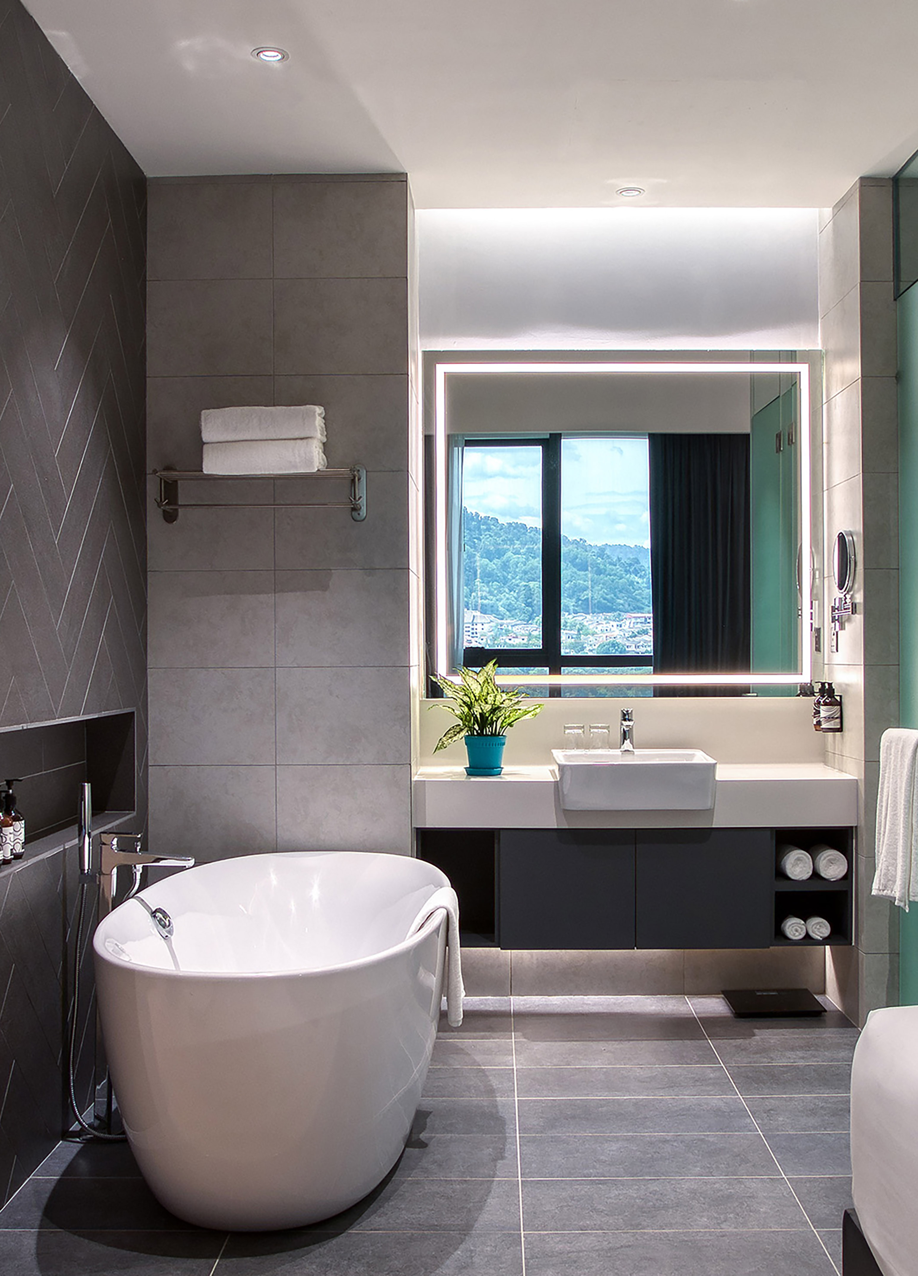 Modern gray tiled bathroom with freestanding tub, vanity, LED mirror, and window view.