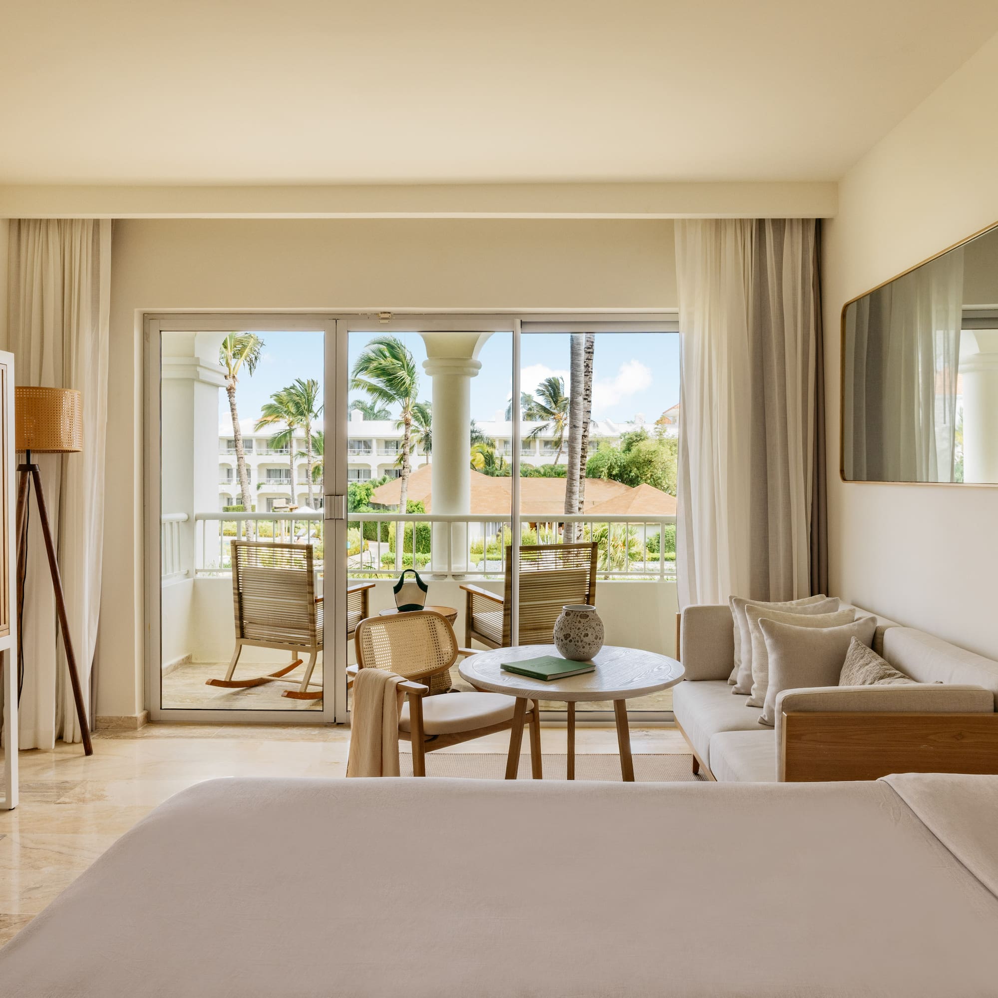 Hotel suite interior with balcony, tropical palm trees, bed, and living area.