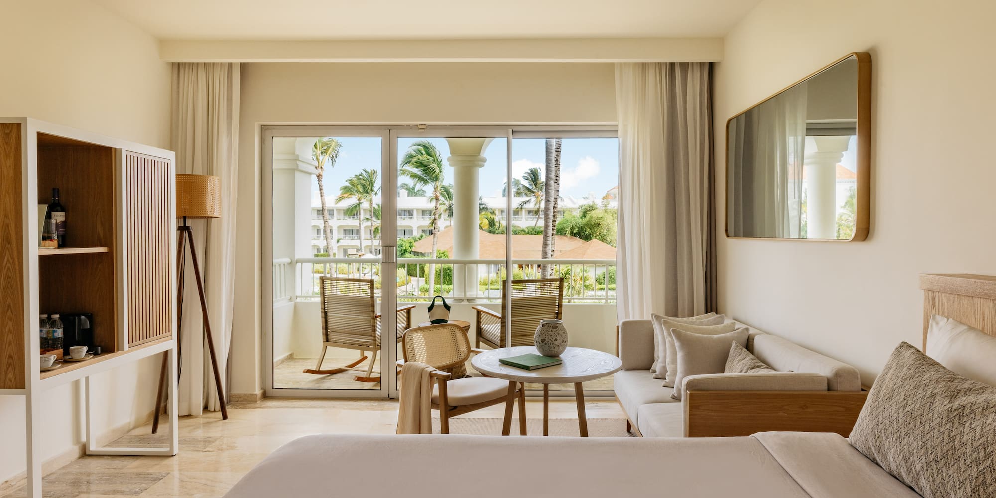 Hotel suite interior with balcony, tropical palm trees, bed, and living area.
