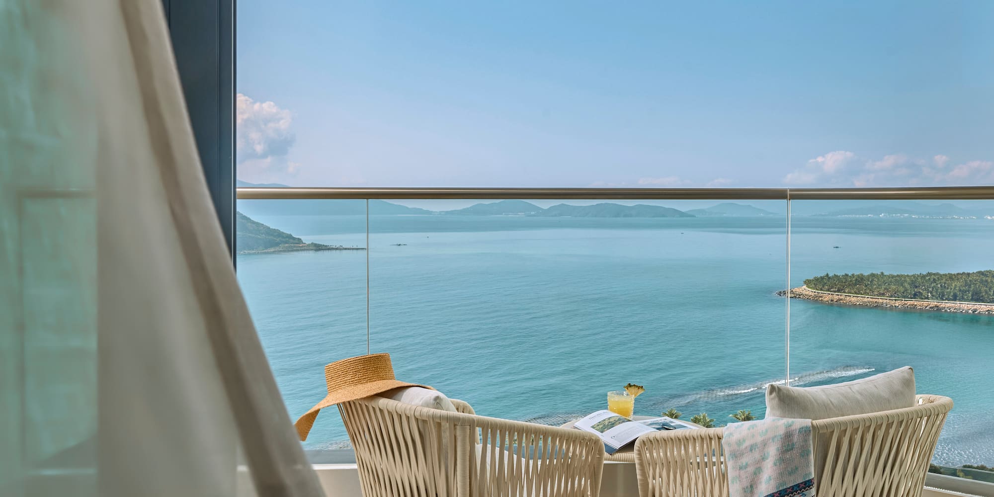 Resort balcony with ocean view, chairs, cocktail, hat, and distant islands.