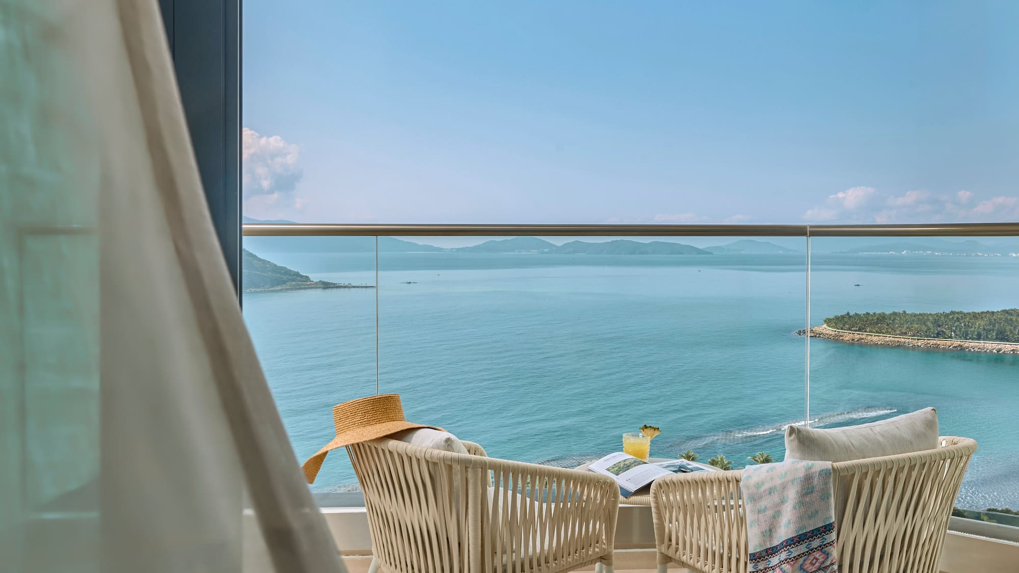 Resort balcony with ocean view, chairs, cocktail, hat, and distant islands.