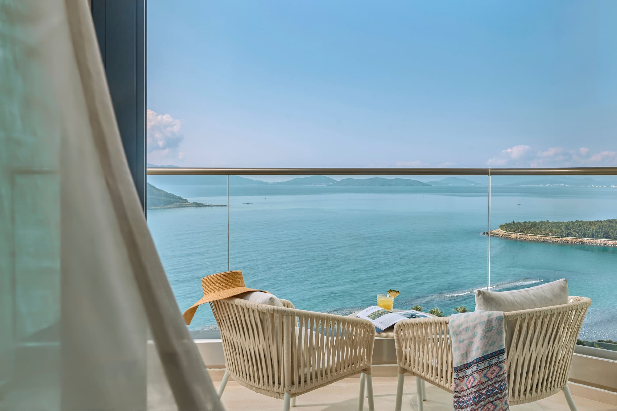 Resort balcony with ocean view, chairs, cocktail, hat, and distant islands.
