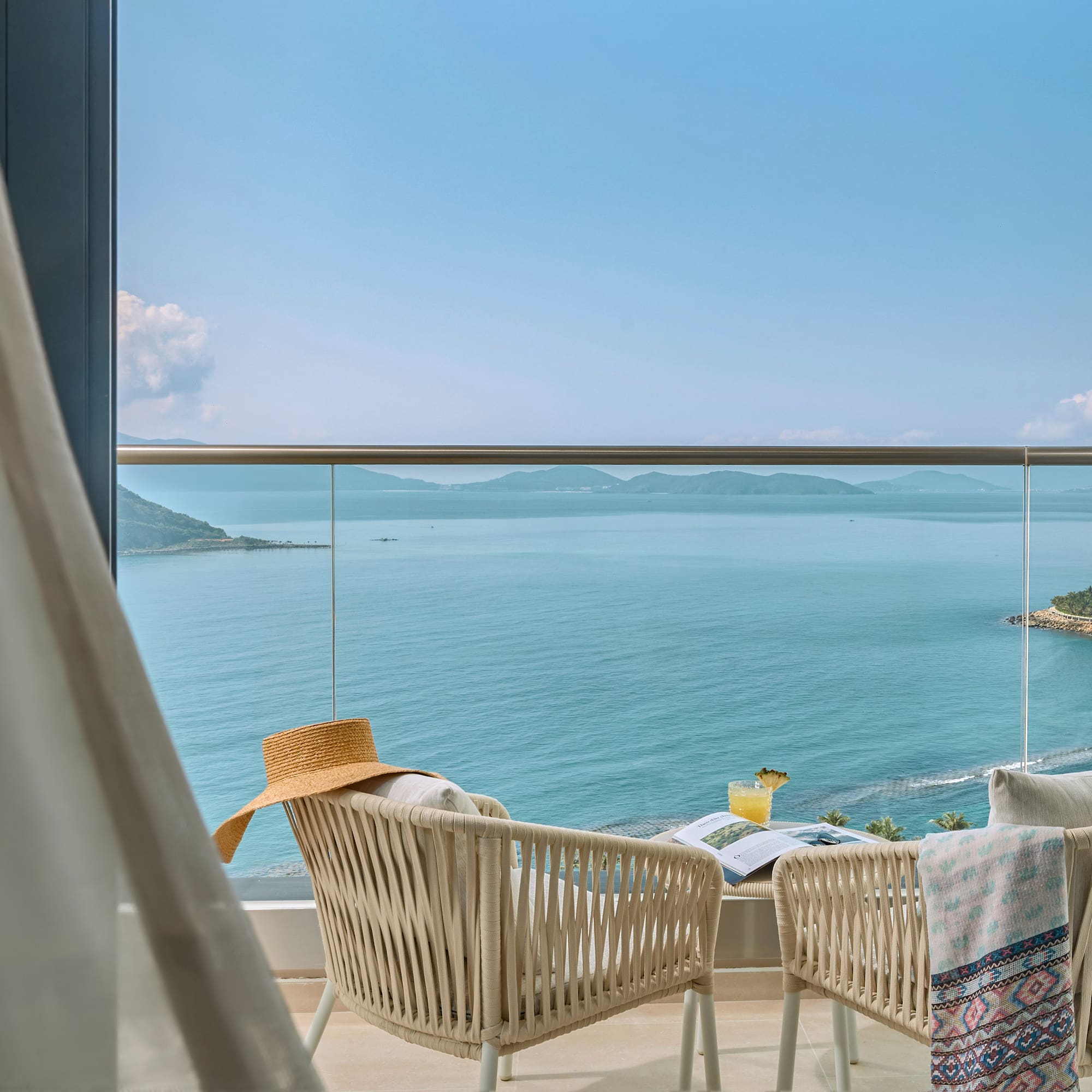Resort balcony with ocean view, chairs, cocktail, hat, and distant islands.