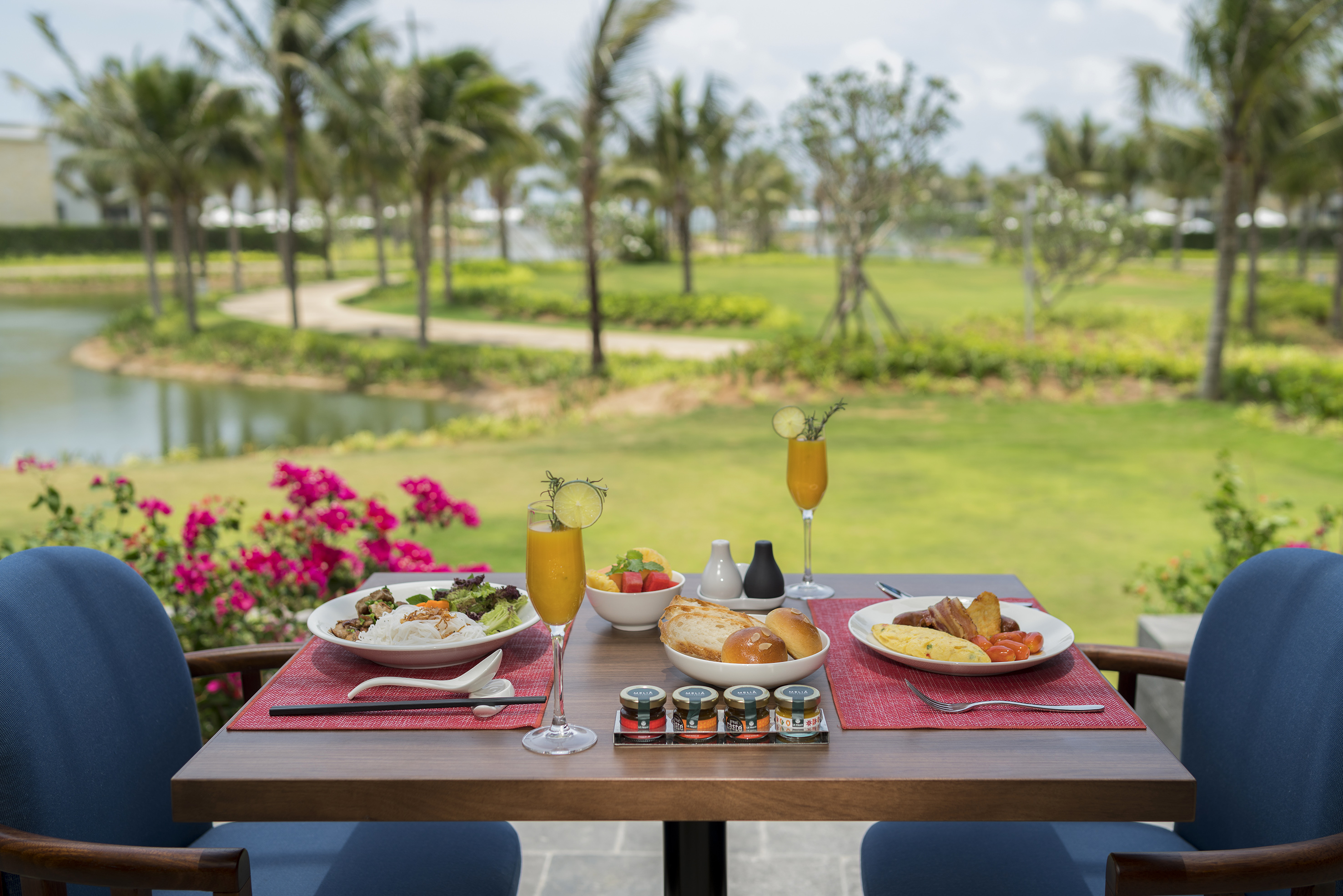 Breakfast spread with mimosas on an outdoor table, palm trees, and lake.