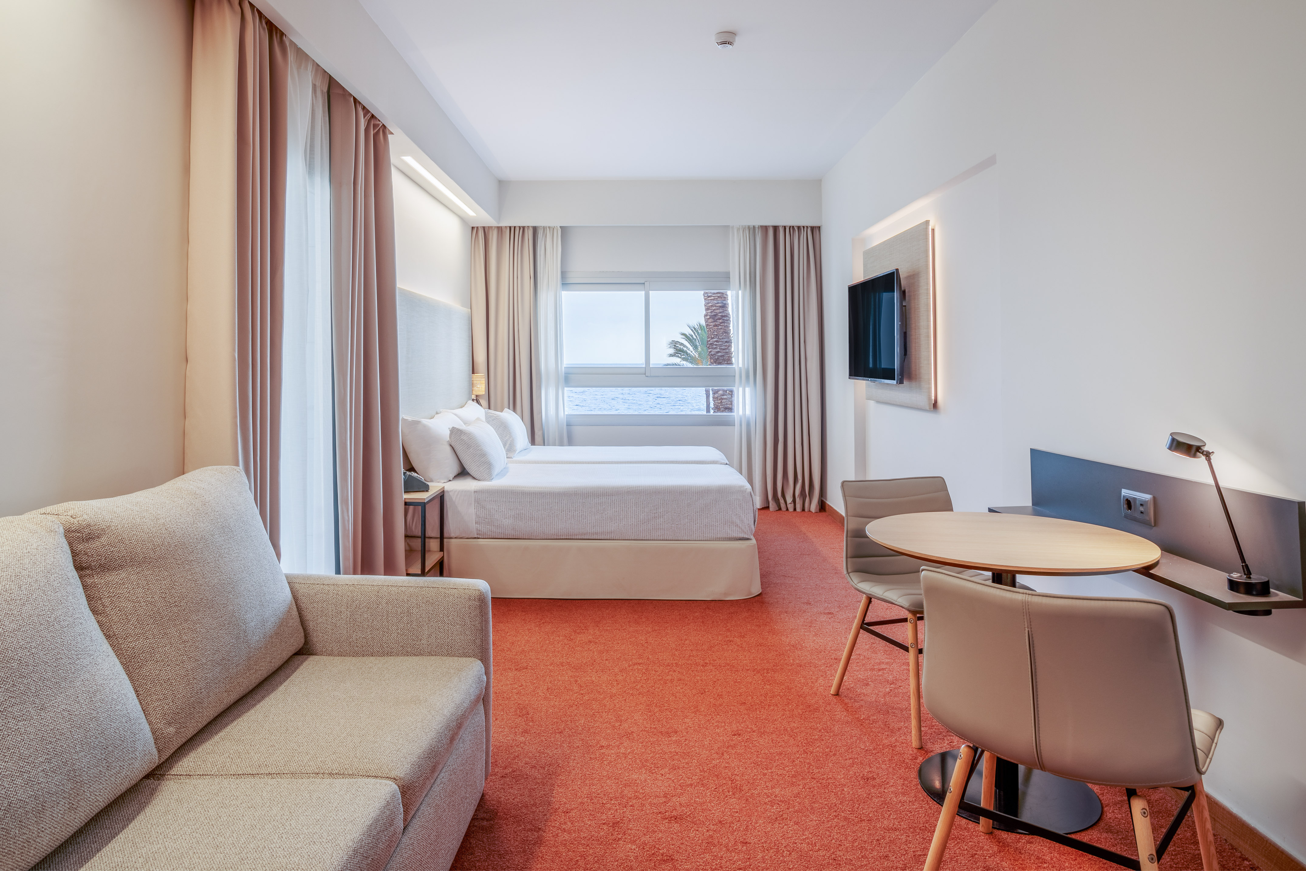 Modern hotel room with twin beds, ocean view, sofa, and dining area.
