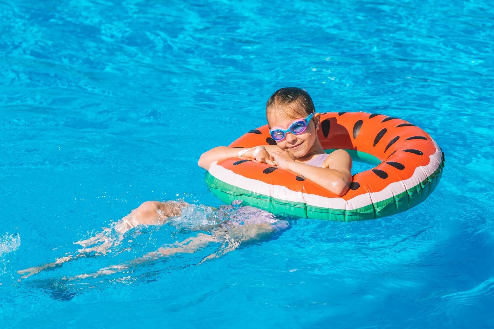 Niña feliz con gafas y flotador de sandía en piscina azul.