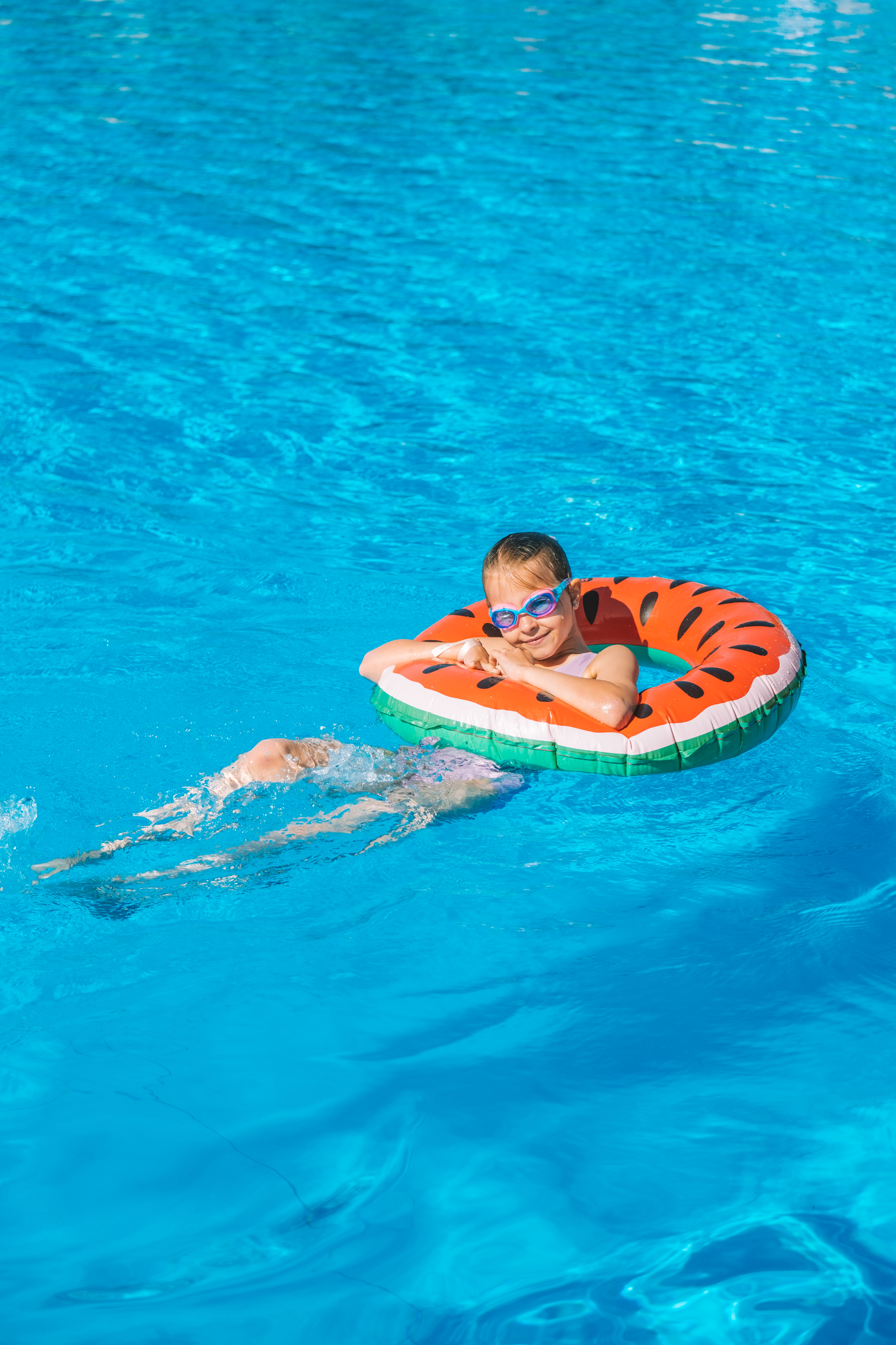 Niña feliz con gafas y flotador de sandía en piscina azul.