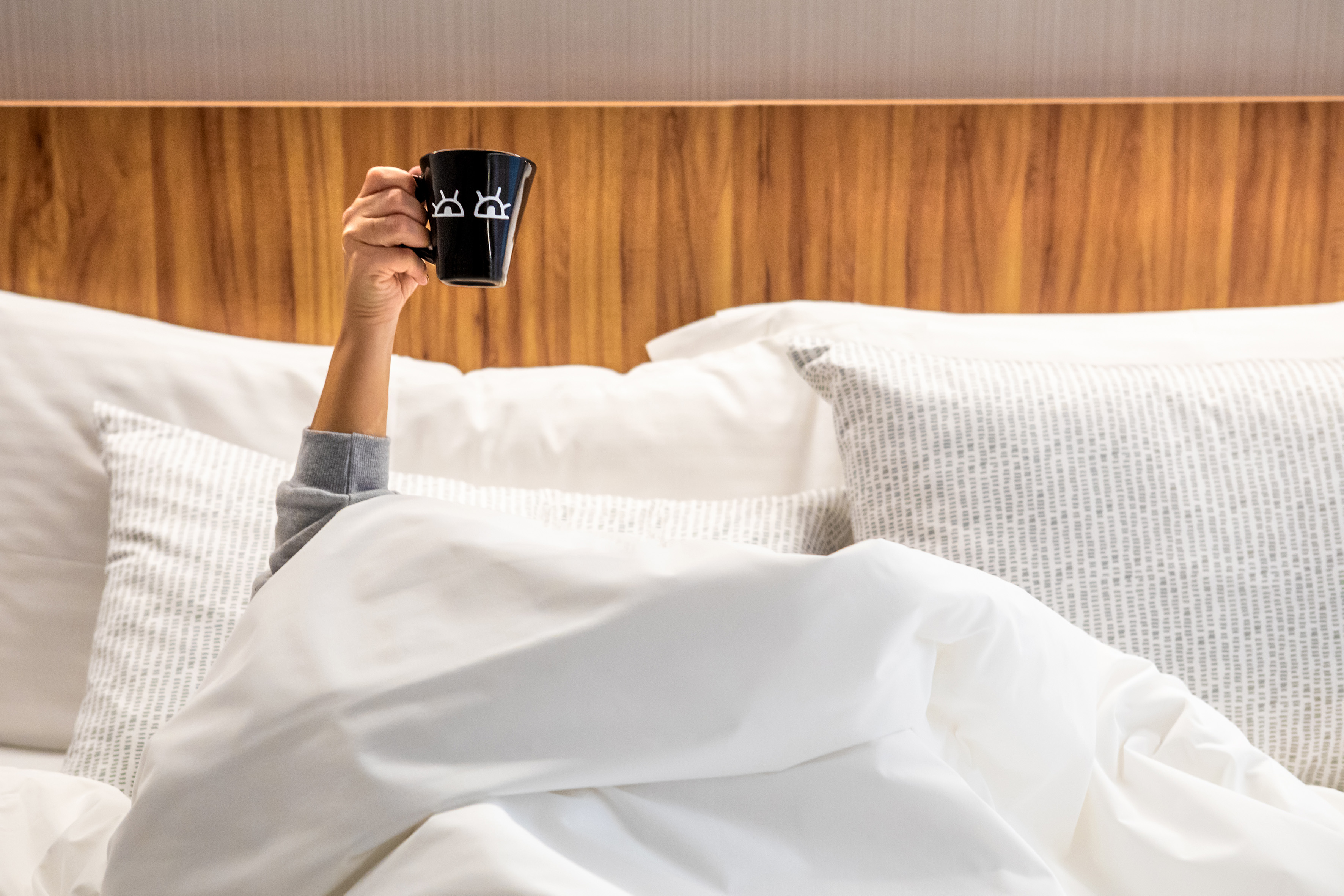 Hand holding black coffee mug with eye icons, emerging from white bed.