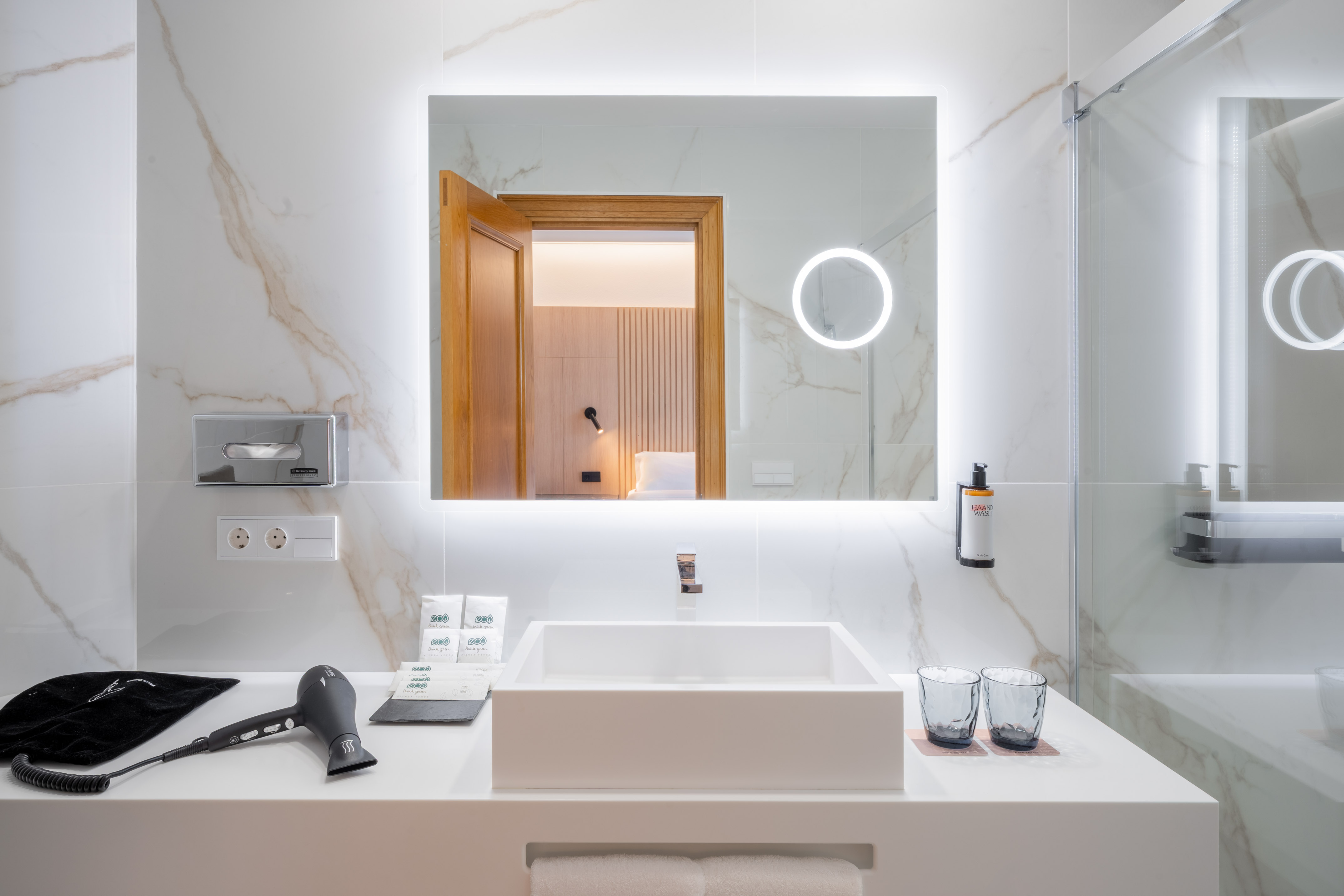 Modern hotel bathroom vanity, backlit mirror, white sink, marble tile walls.