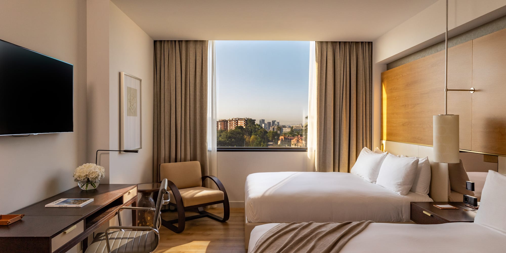 Hotel room with two beds, work desk, and city view window.