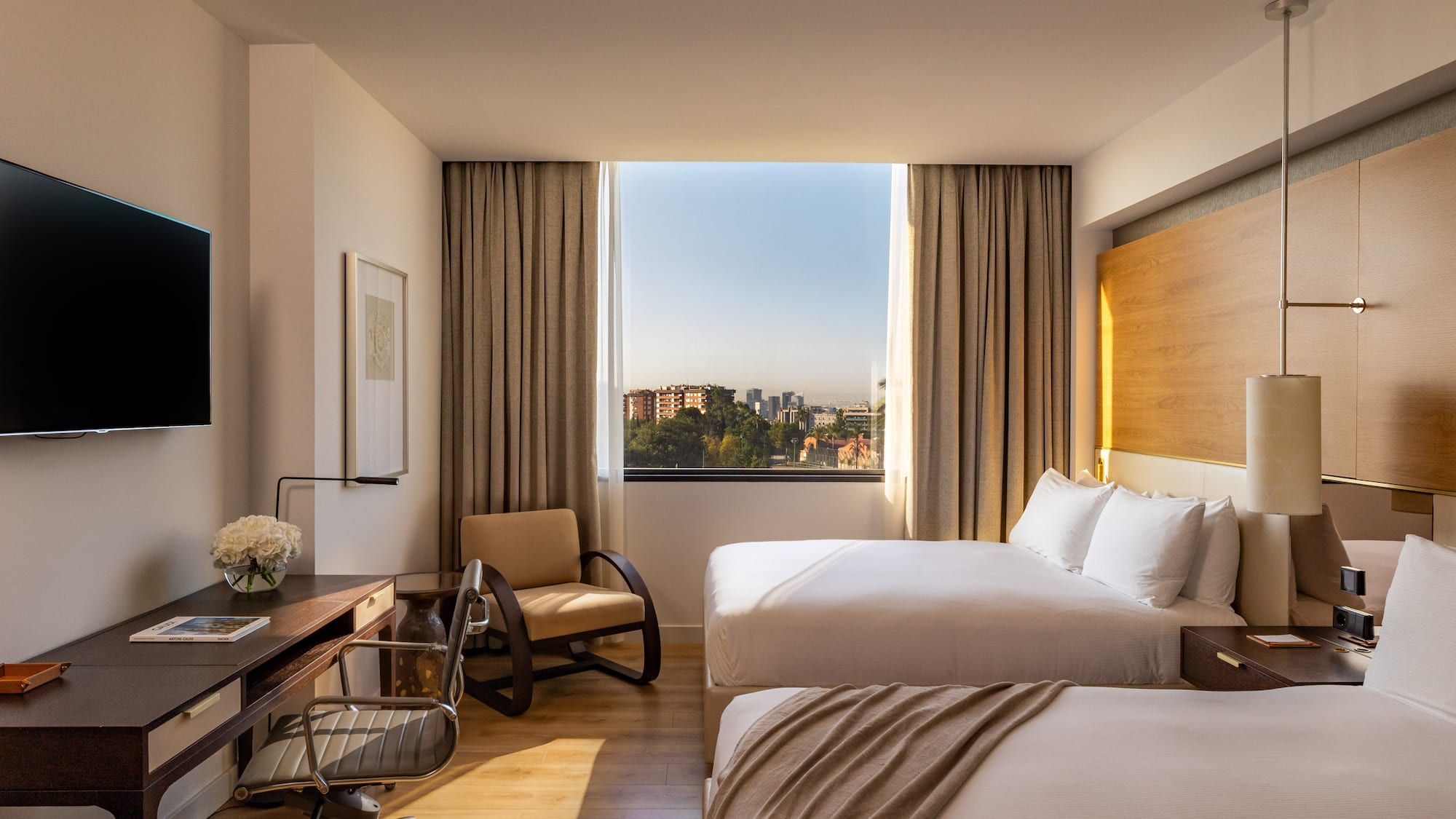 Hotel room with two beds, work desk, and city view window.