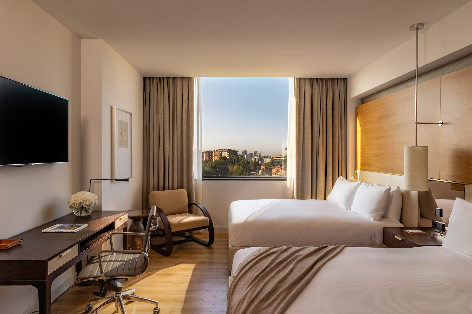 Hotel room with two beds, work desk, and city view window.