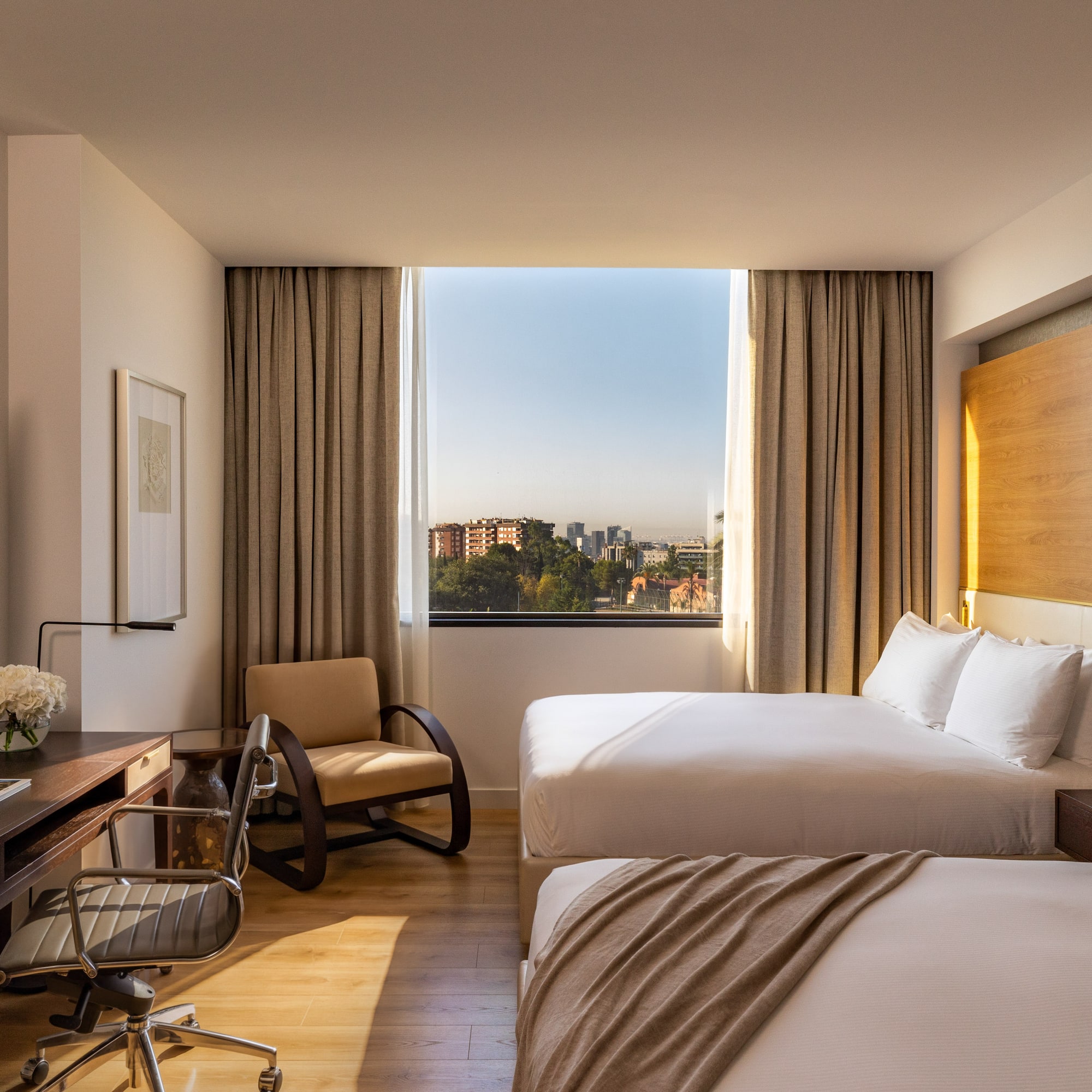 Hotel room with two beds, work desk, and city view window.