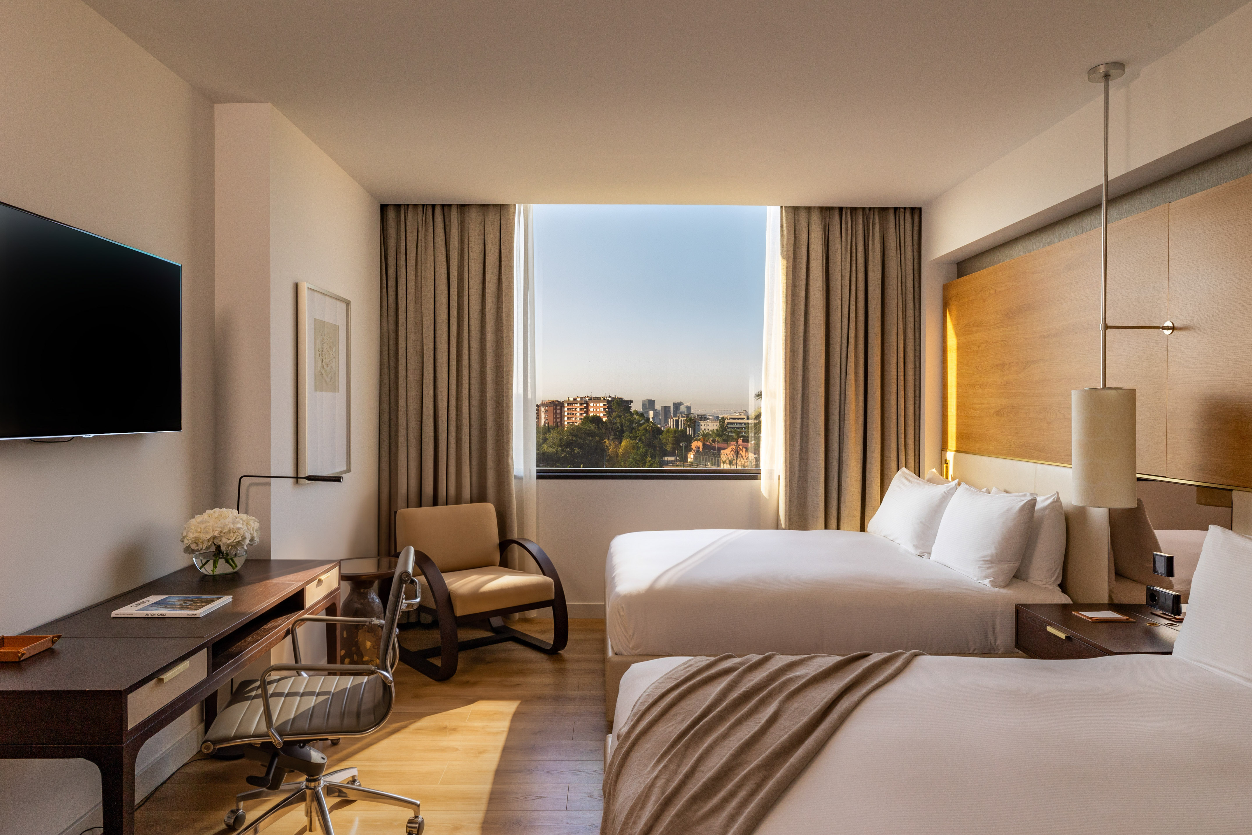 Hotel room with two beds, work desk, and city view window.