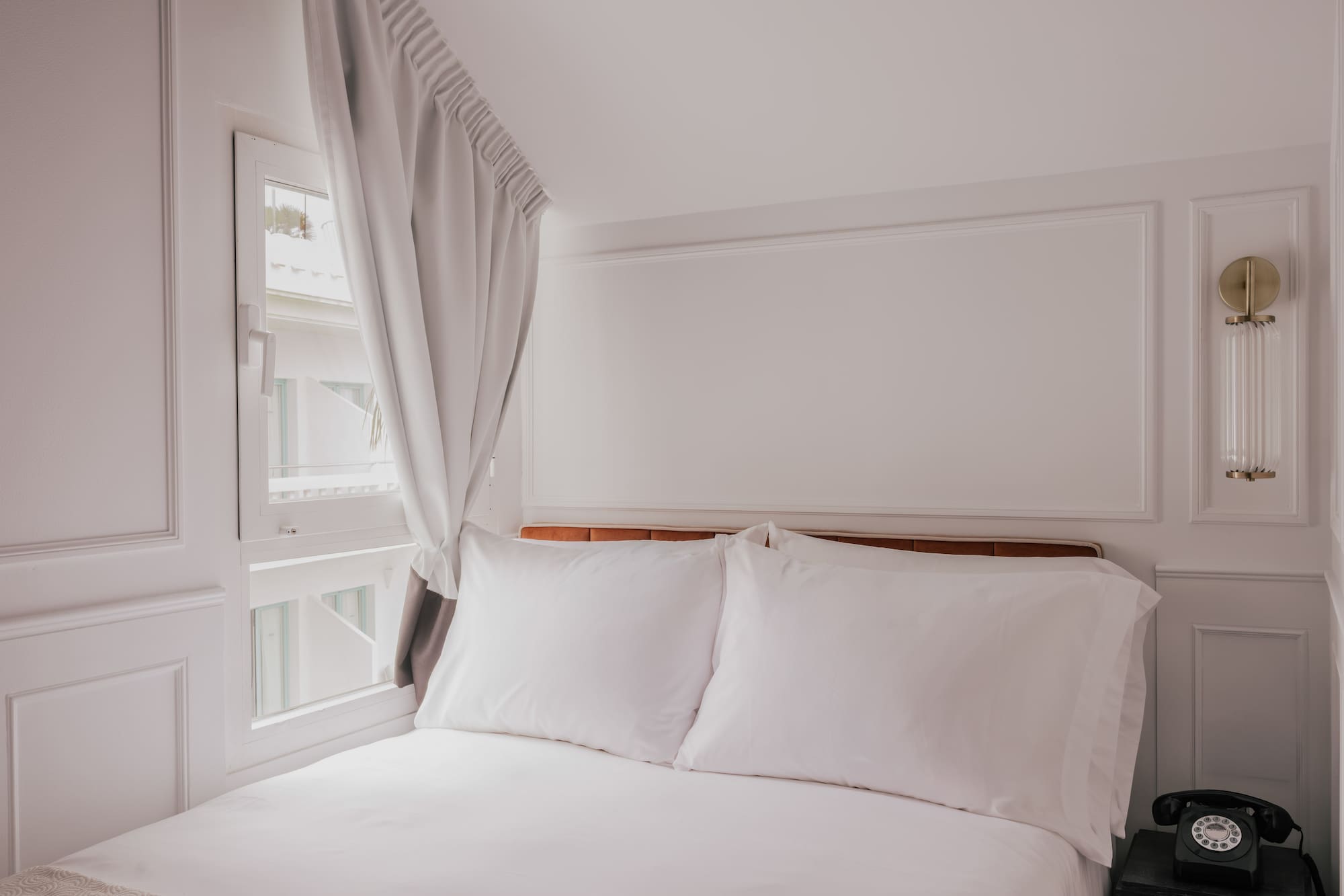 Clean white hotel bedroom with bed, window, nightstand, and vintage phone.