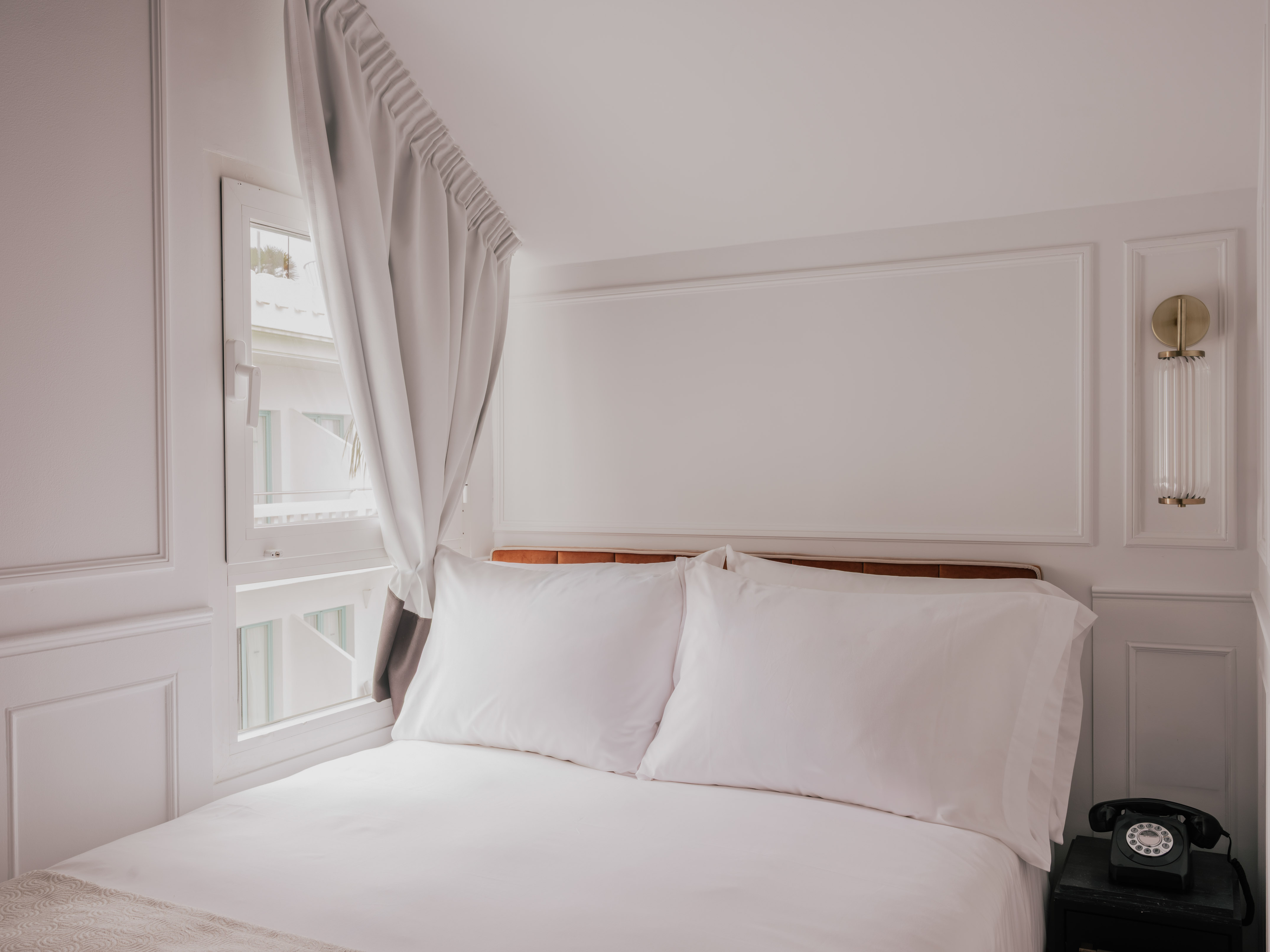 Clean white hotel bedroom with bed, window, nightstand, and vintage phone.