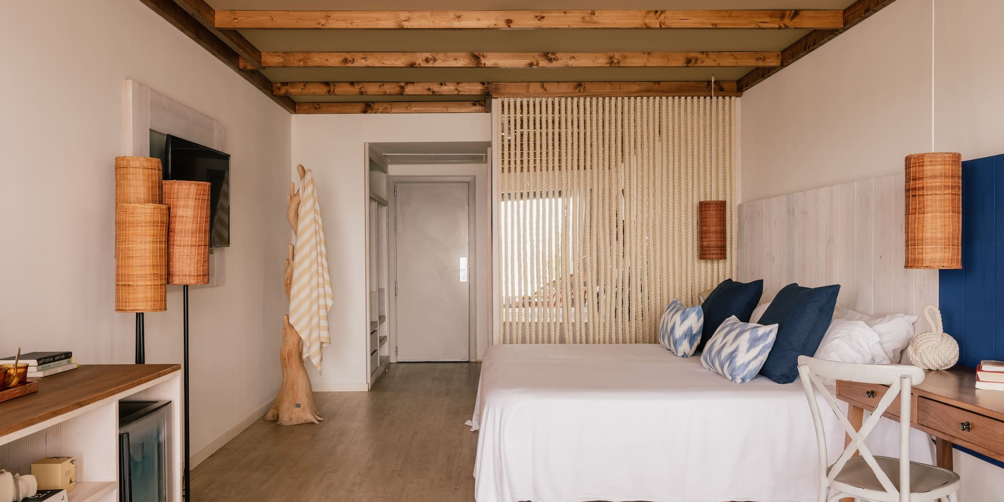 Dormitorio de hotel boho con cama blanca, vigas de madera y separador de cuerda.