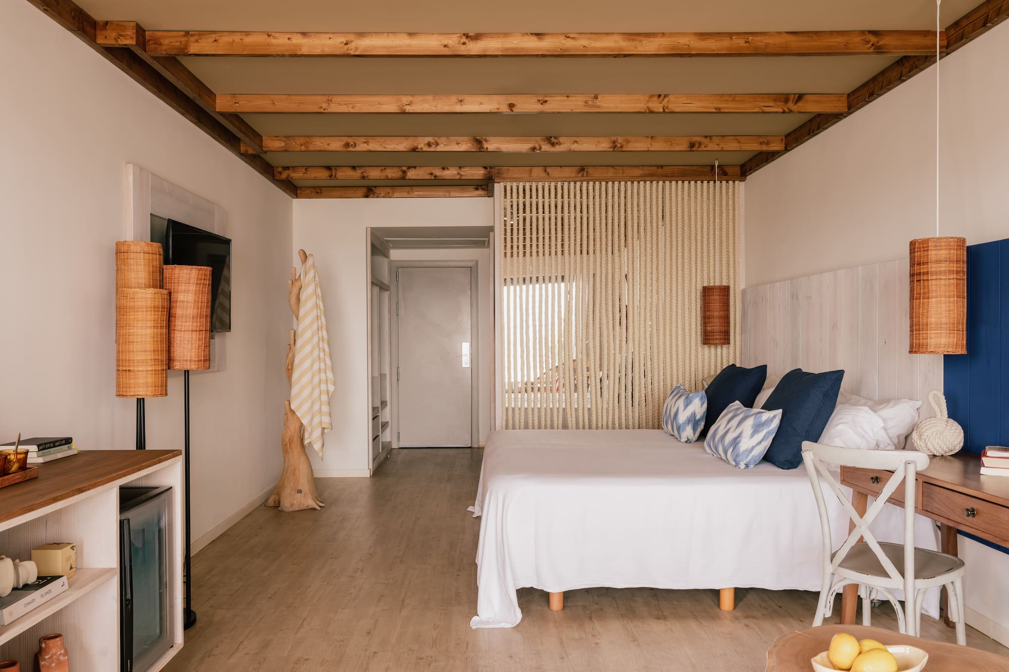 Dormitorio de hotel boho con cama blanca, vigas de madera y separador de cuerda.