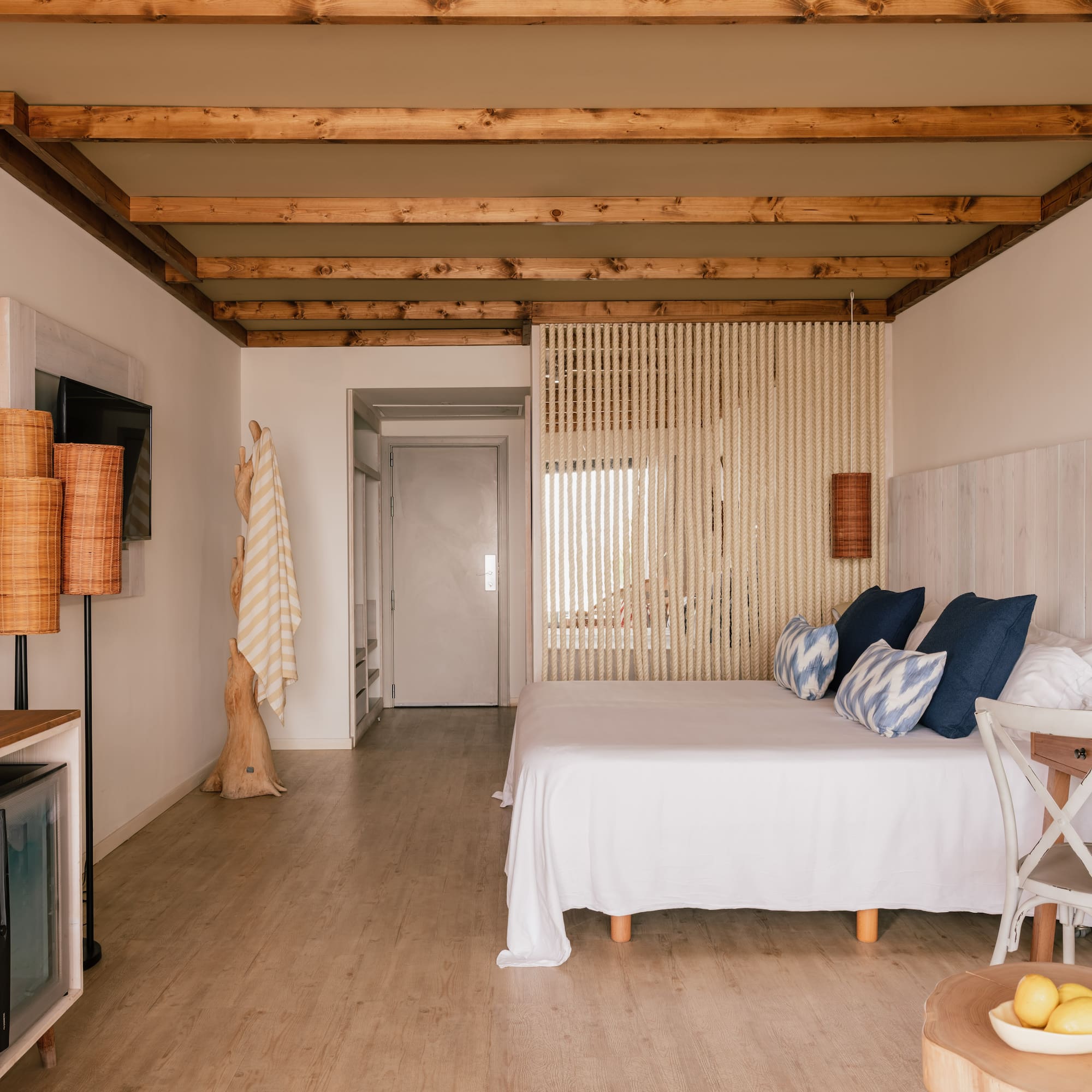 Dormitorio de hotel boho con cama blanca, vigas de madera y separador de cuerda.