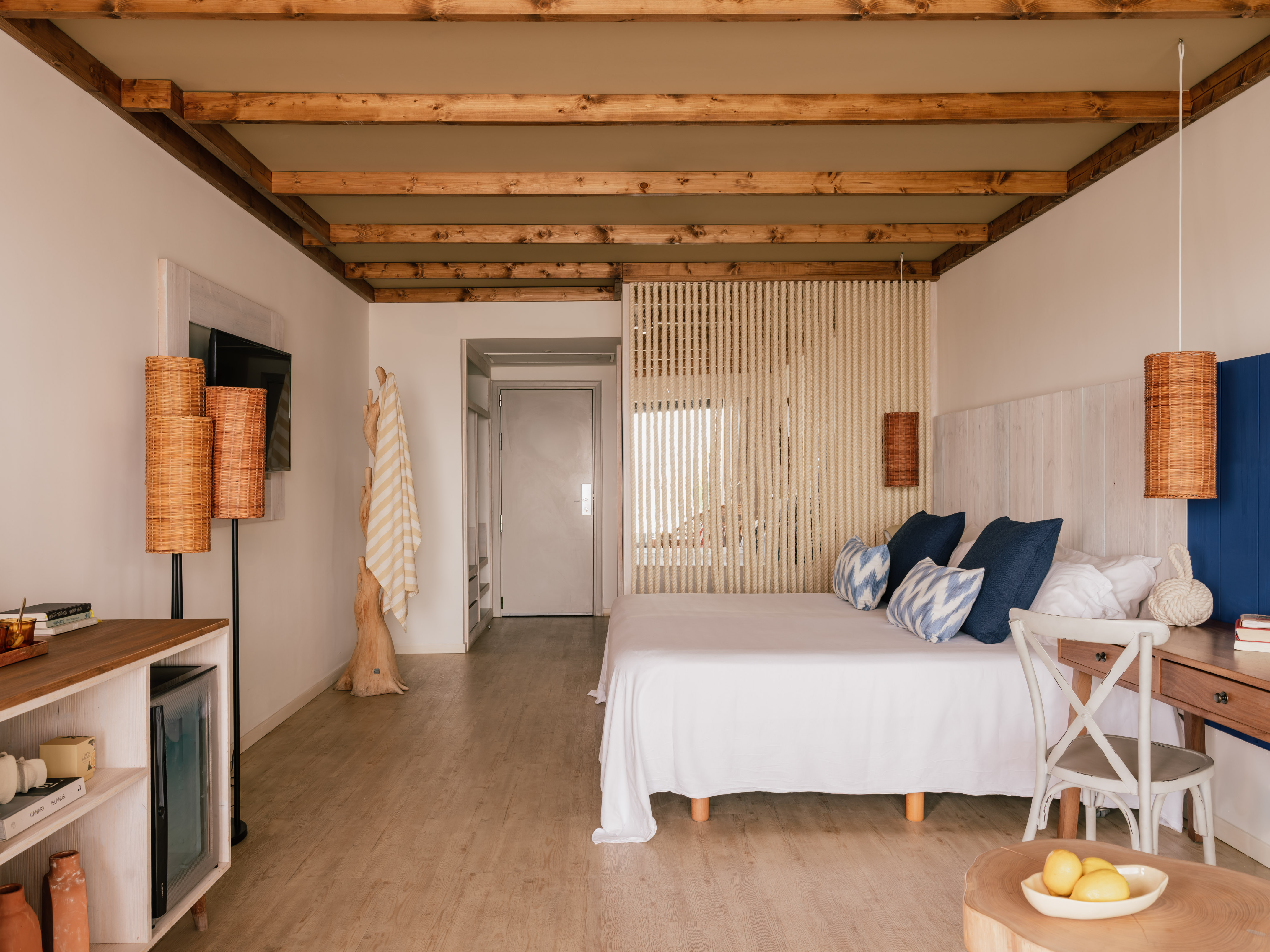 Dormitorio de hotel boho con cama blanca, vigas de madera y separador de cuerda.