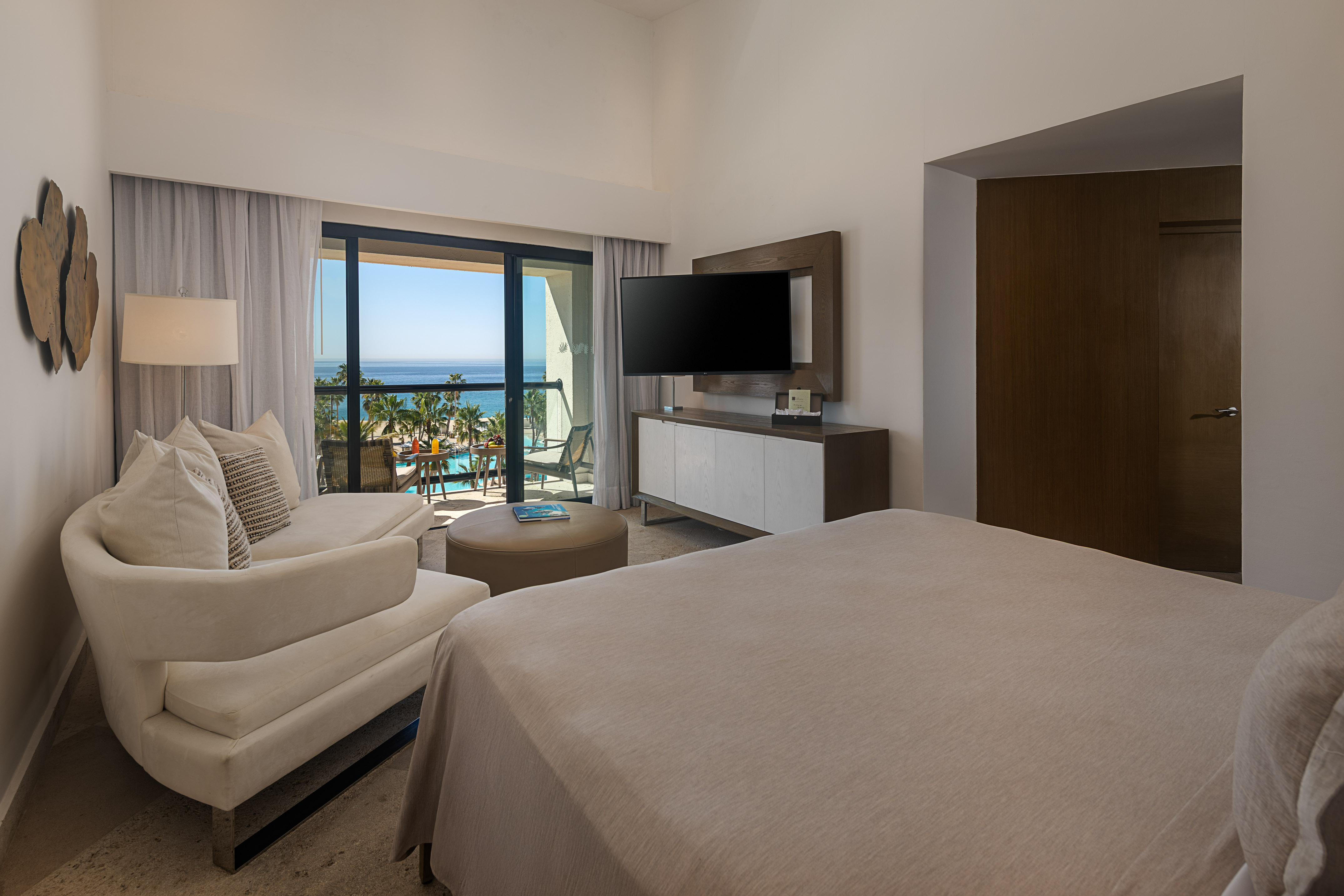 Modern hotel room with ocean view balcony, bed, sofa, and tropical resort pool.