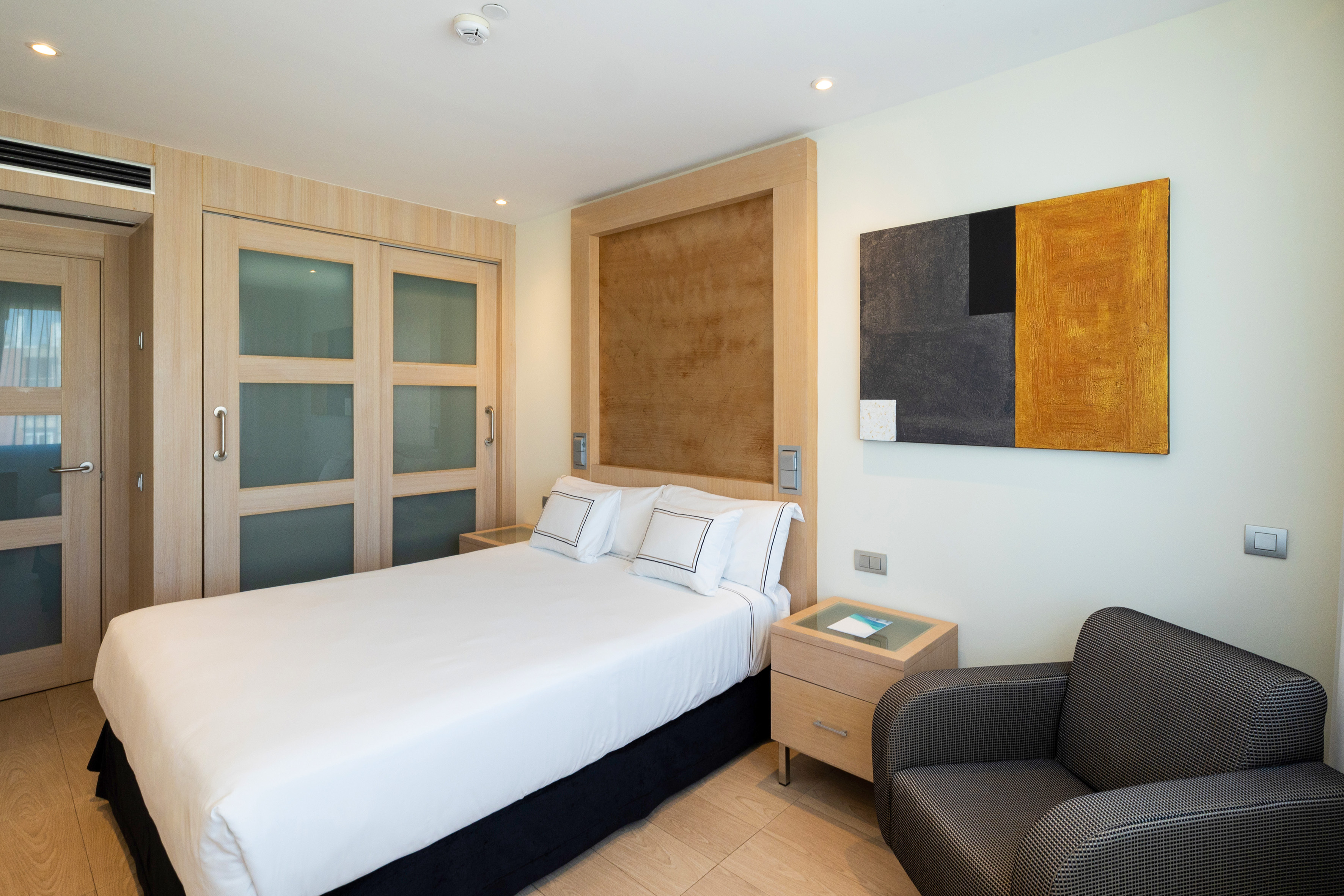 Modern hotel room, white bed, light wood furniture, abstract art.