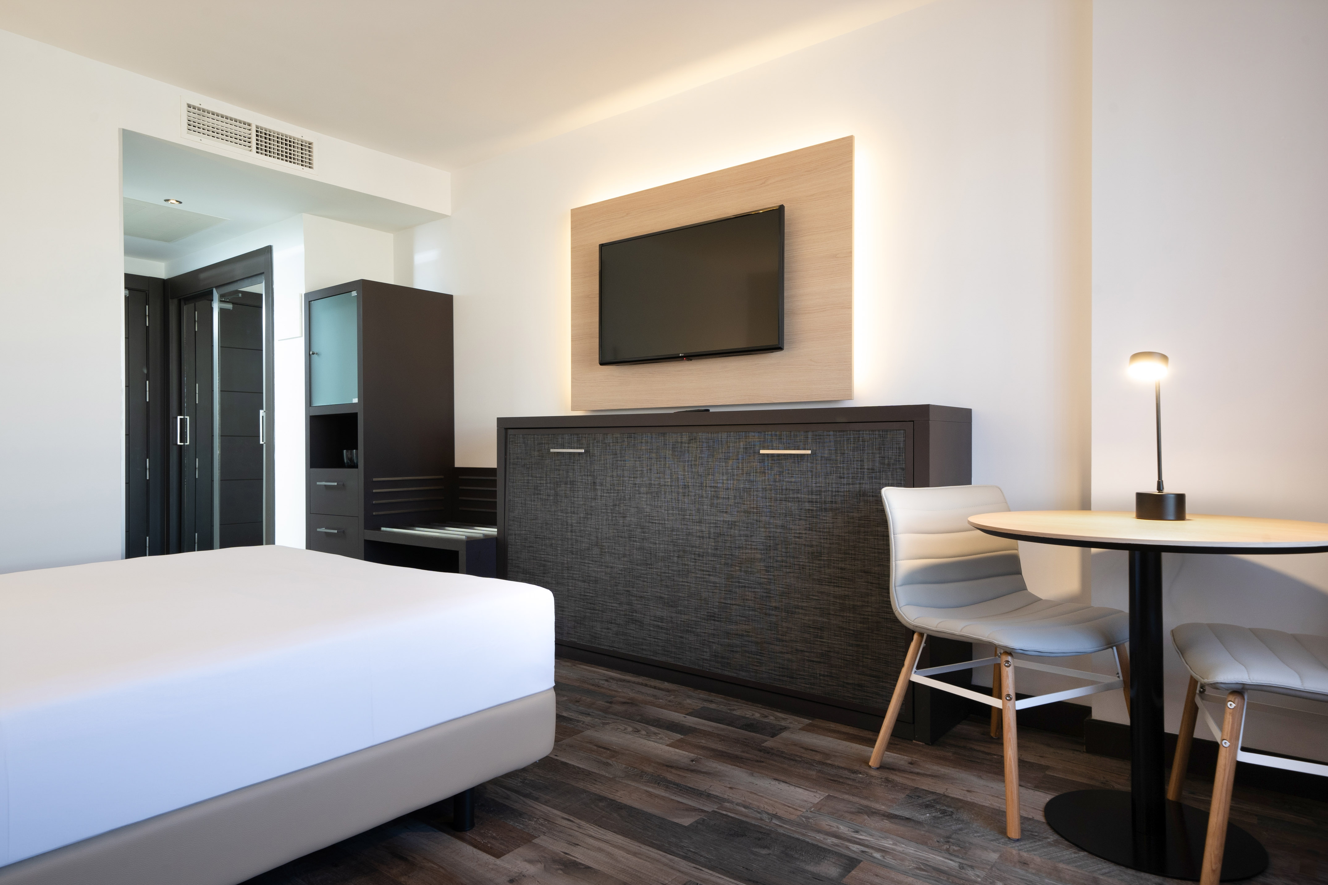Modern hotel room: bed, wall TV, murphy bed unit, small dining table.