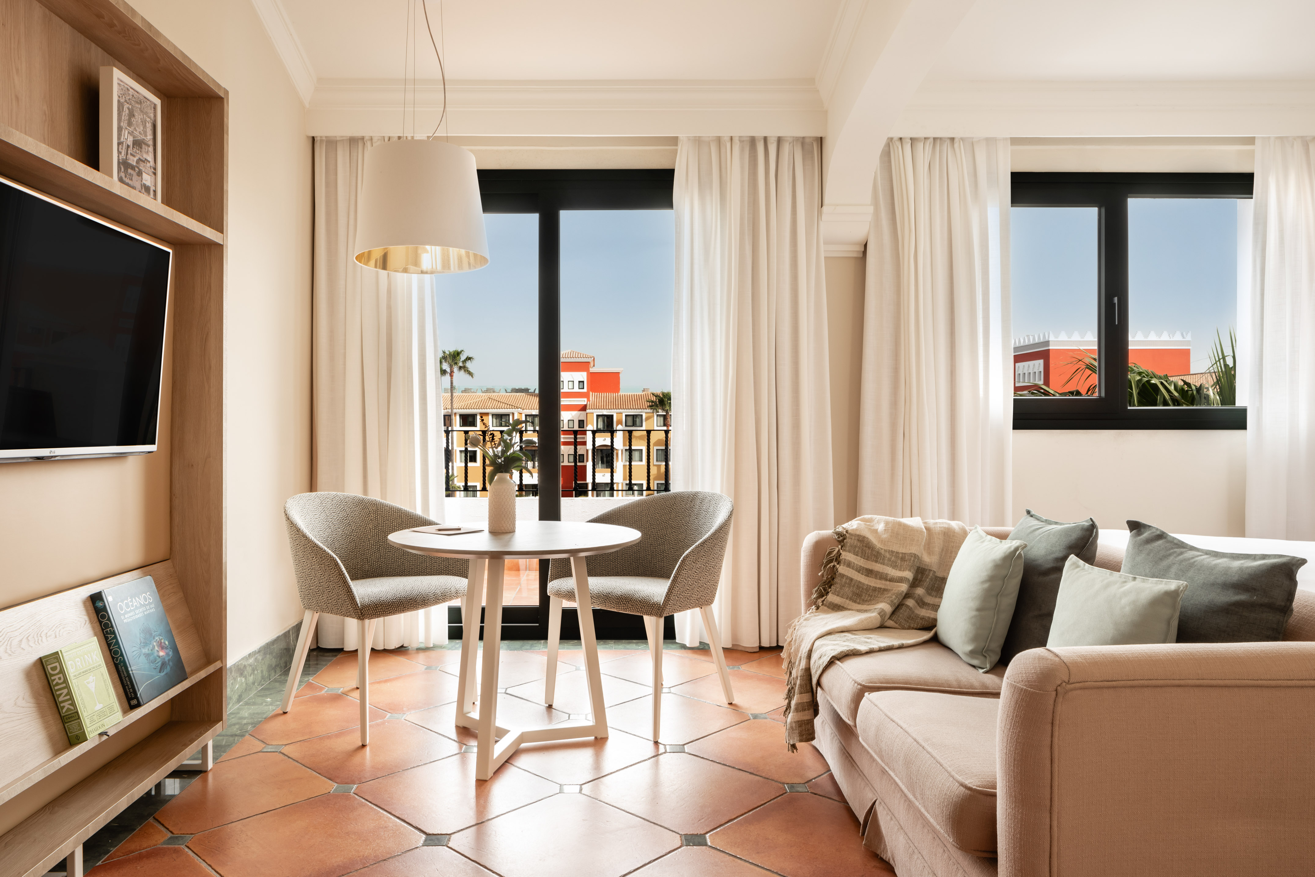 Hotel suite with sofa, dining table, TV, and balcony overlooking resort.