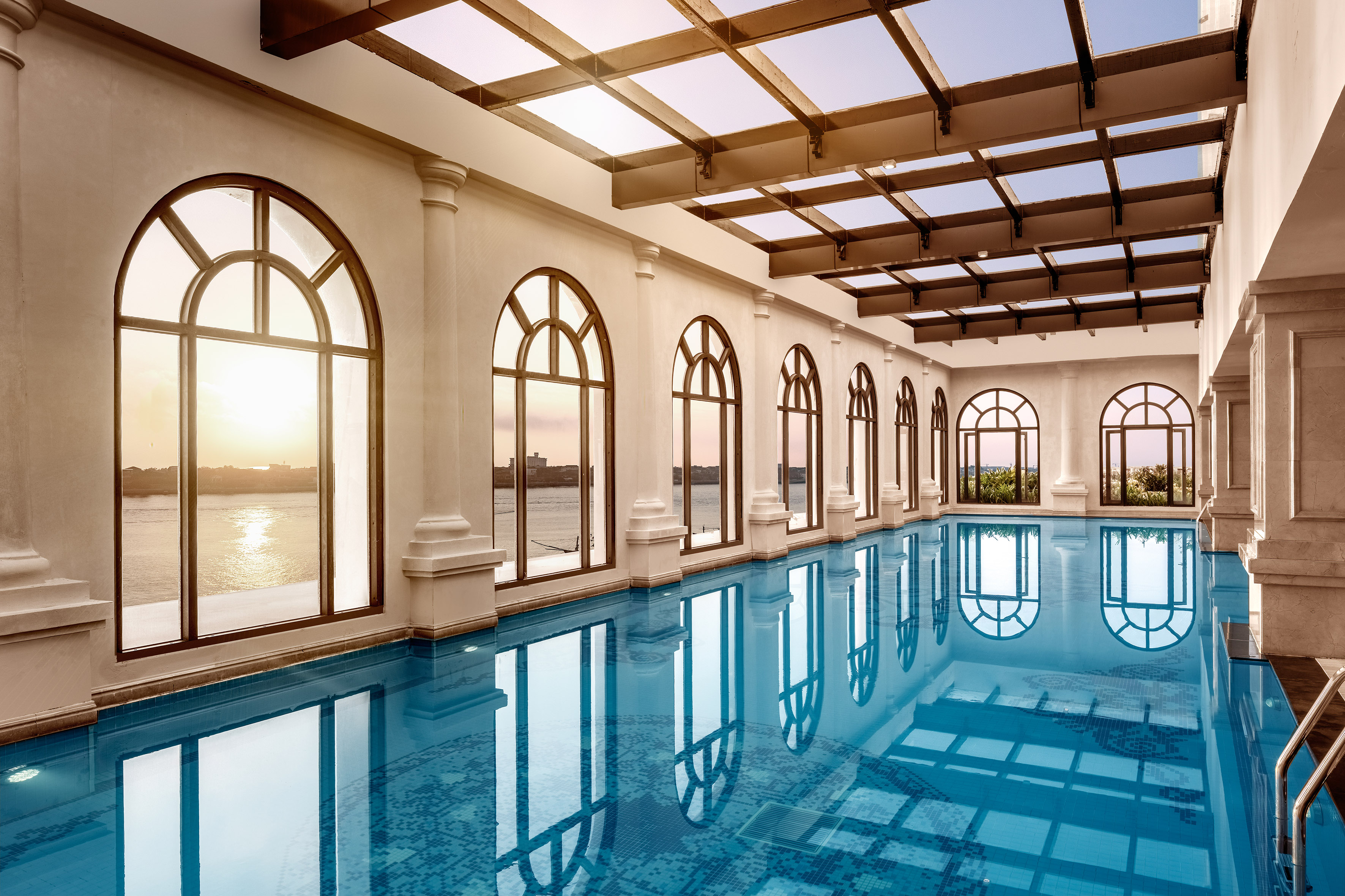 Indoor swimming pool with large arched windows overlooking water at sunset.
