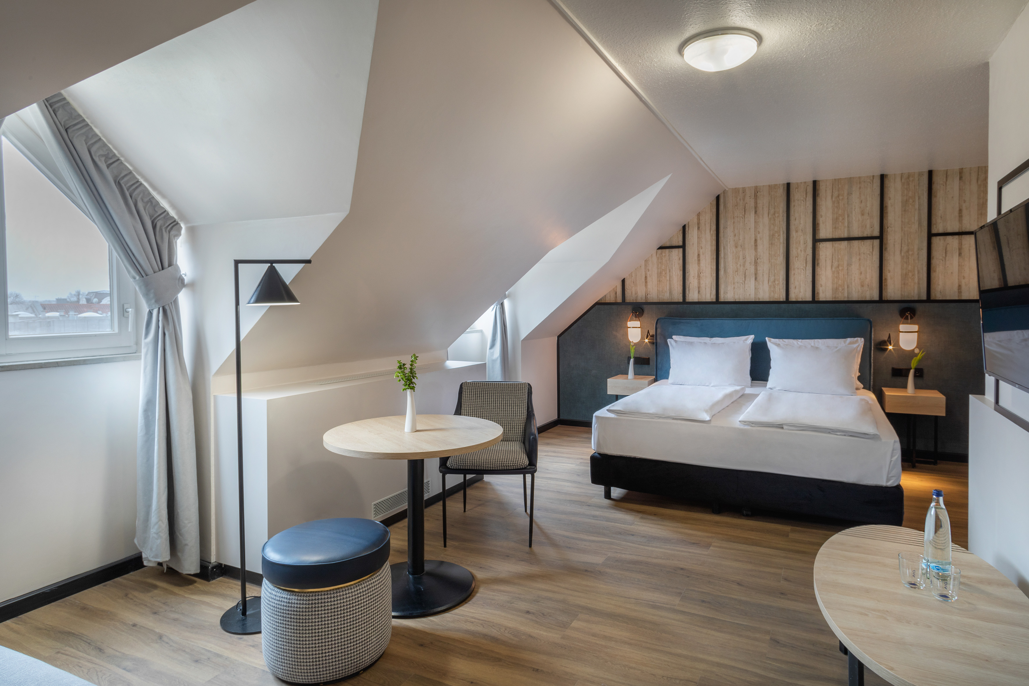 Modern attic hotel room with bed, table, and wood floor.