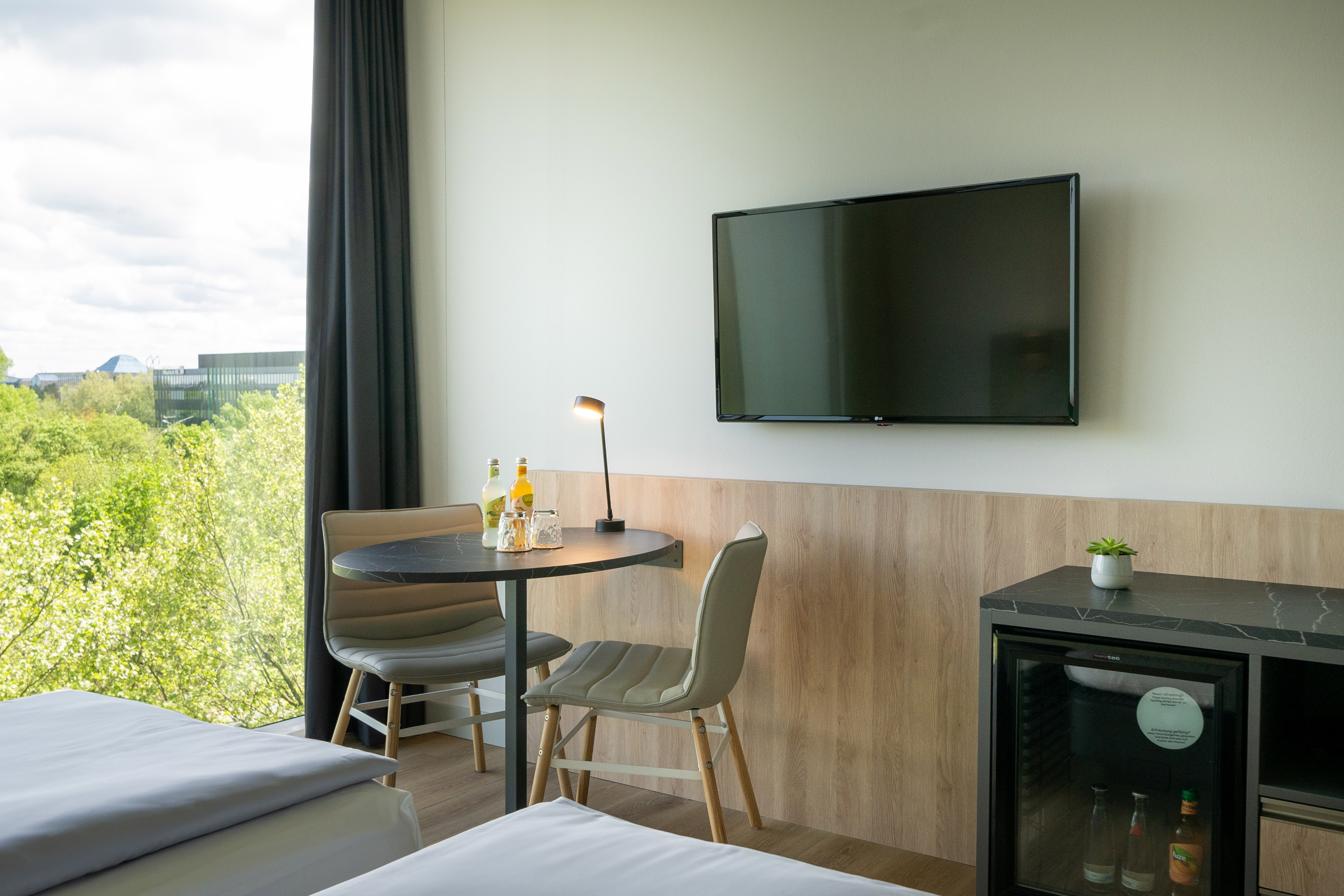 Modern hotel room with city view, dining area, TV, and mini-fridge.