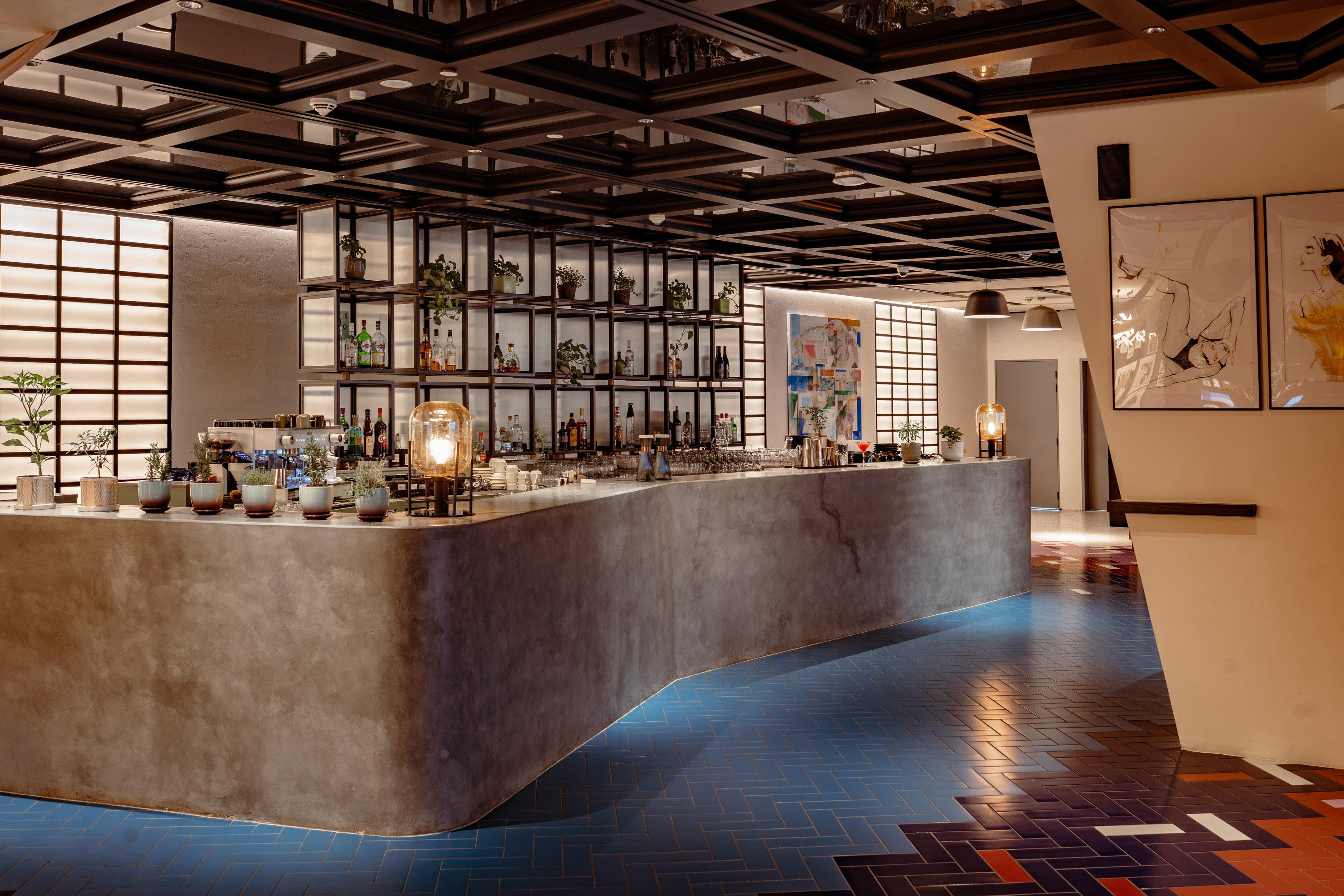 Modern hotel bar with concrete counter, backlit shelves, and herringbone floor.