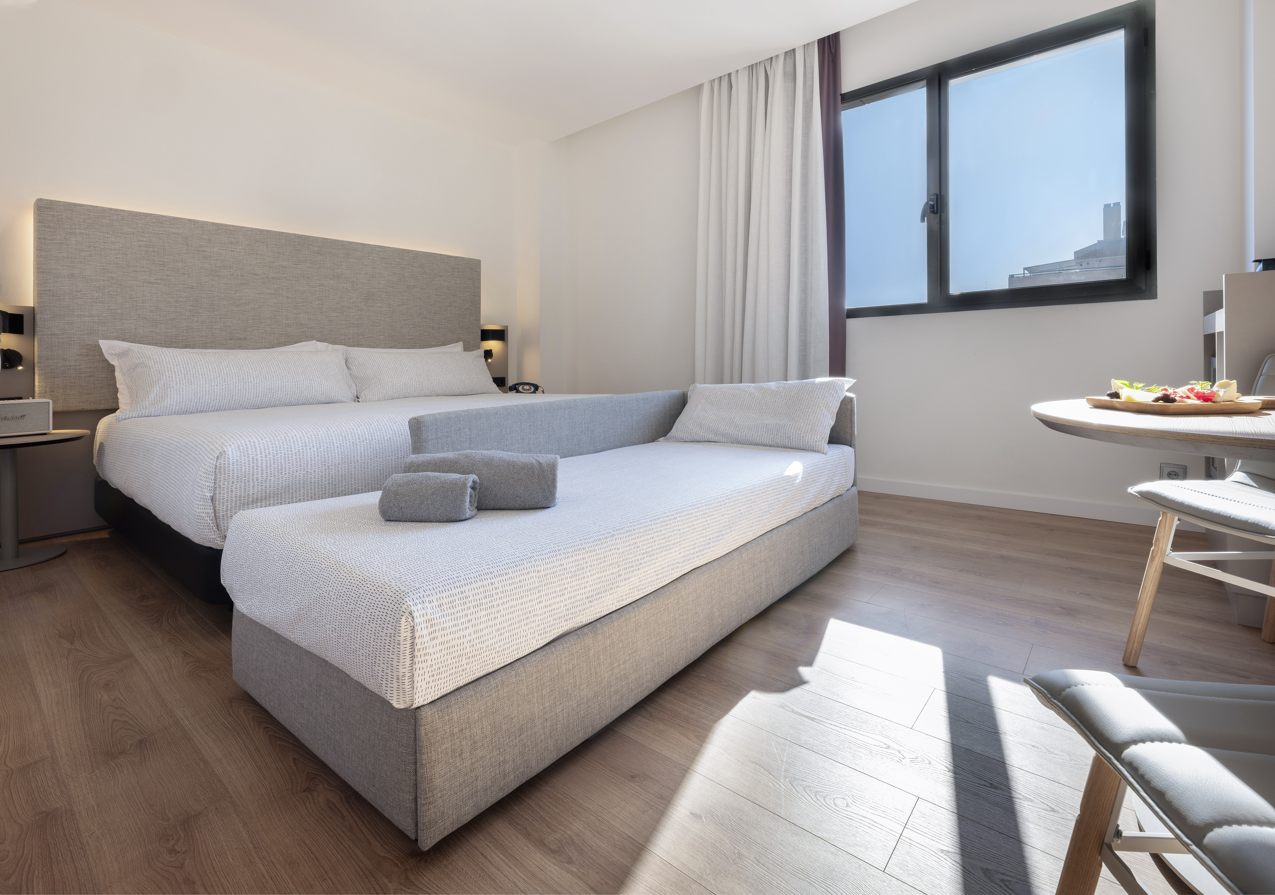 Modern hotel bedroom with bed, daybed, wood floor, and bright window.