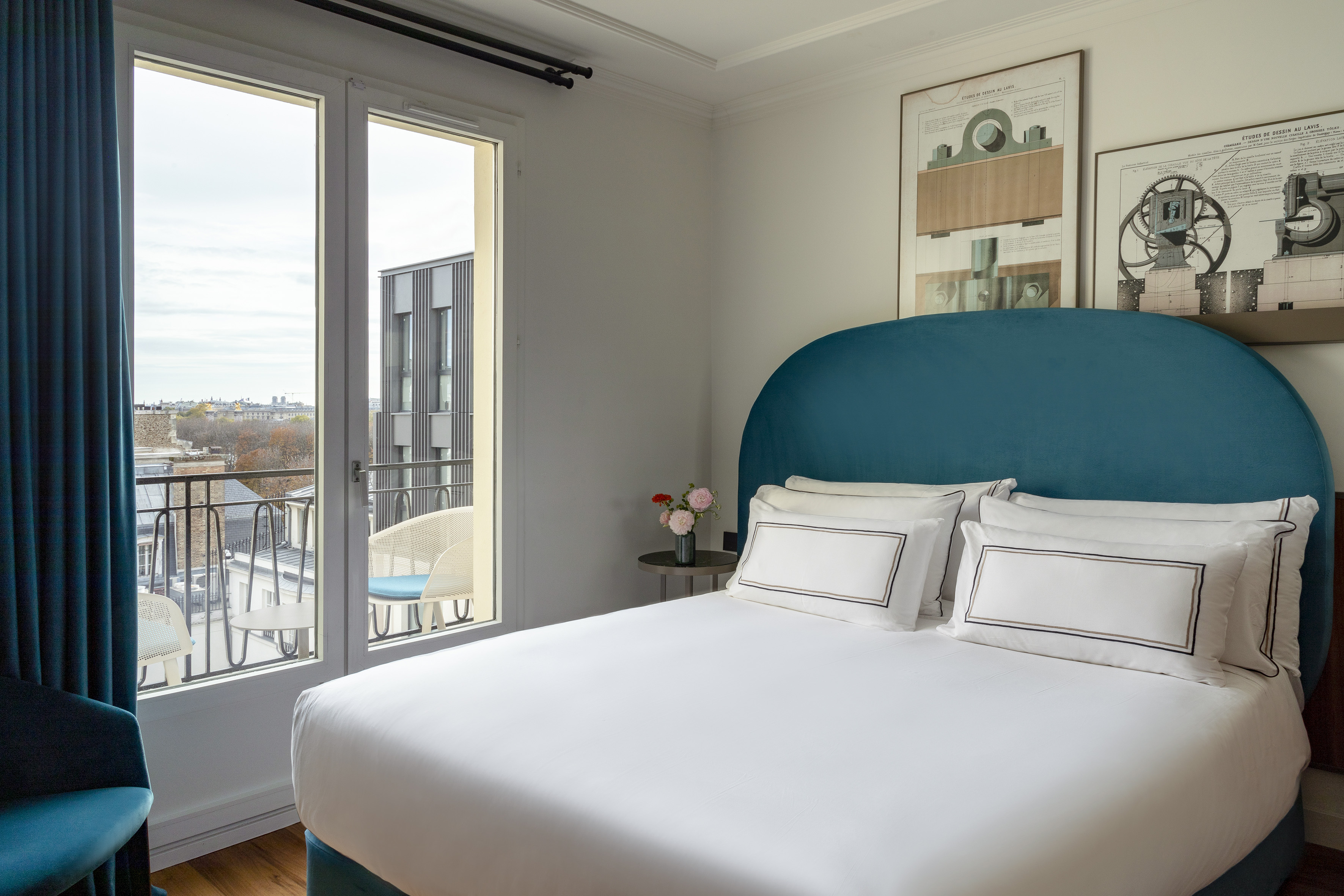 Modern hotel bedroom with white bed, teal headboard, balcony, and city view.