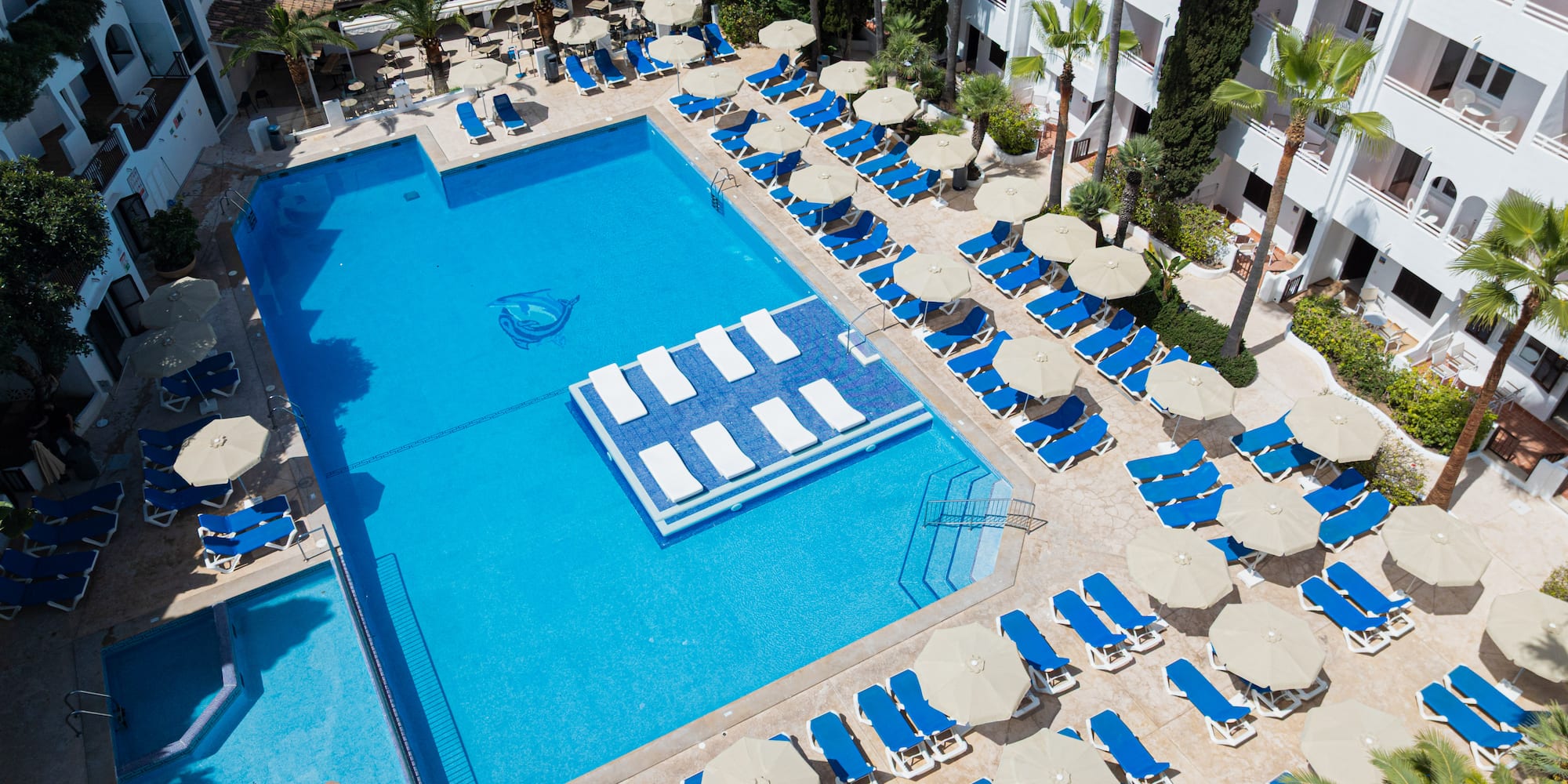 Aerial view of a resort pool with blue loungers, umbrellas, and palm trees.
